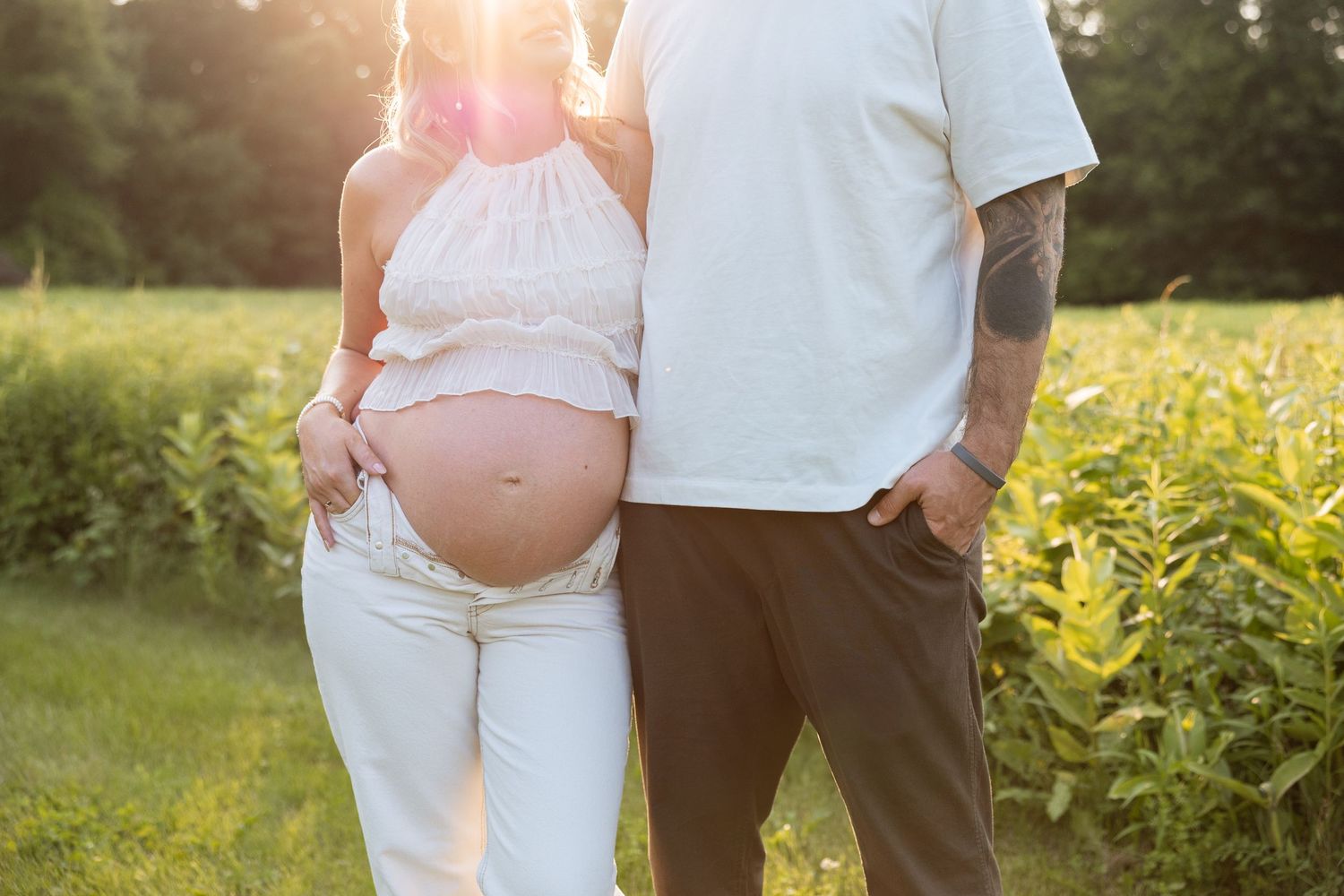 A couple stands in a sunlit field during a maternity photo shoot, with the pregnant belly illuminated by golden hour light.