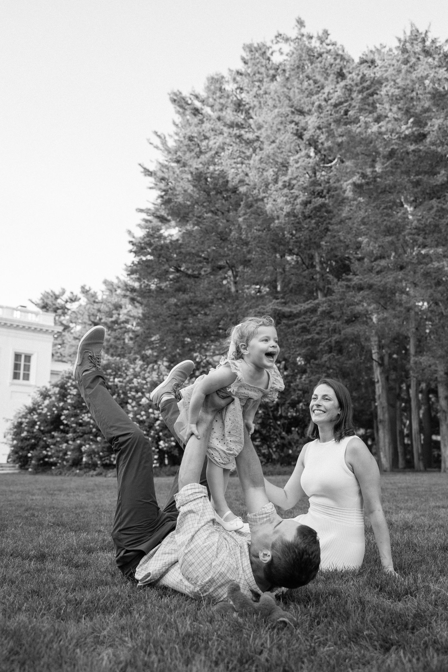 A joyful family plays together outdoors on a lawn with trees in the background.