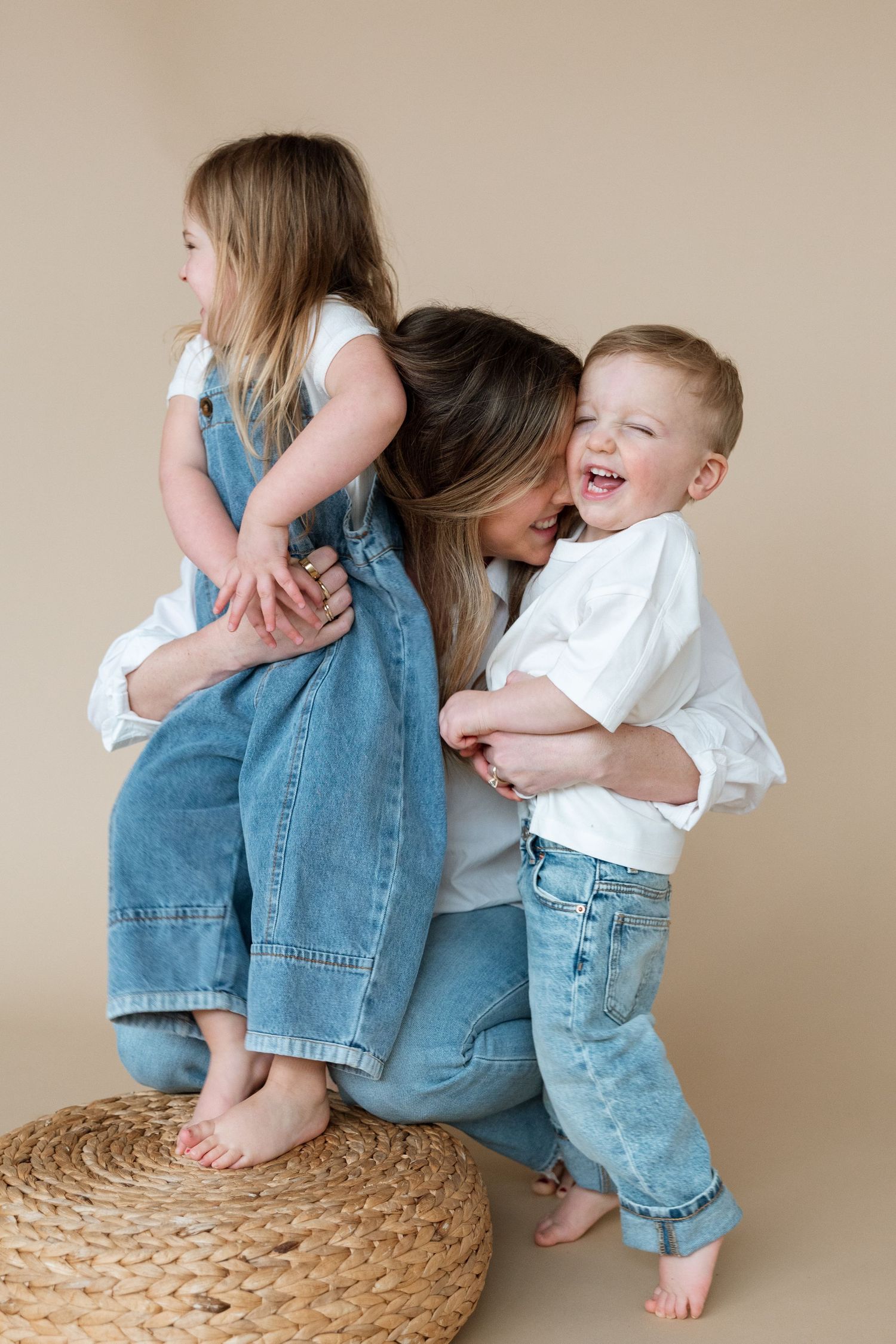 Sweet candid moments of parent and child laughing and playing while sitting on a woven ottoman against beige wall.
