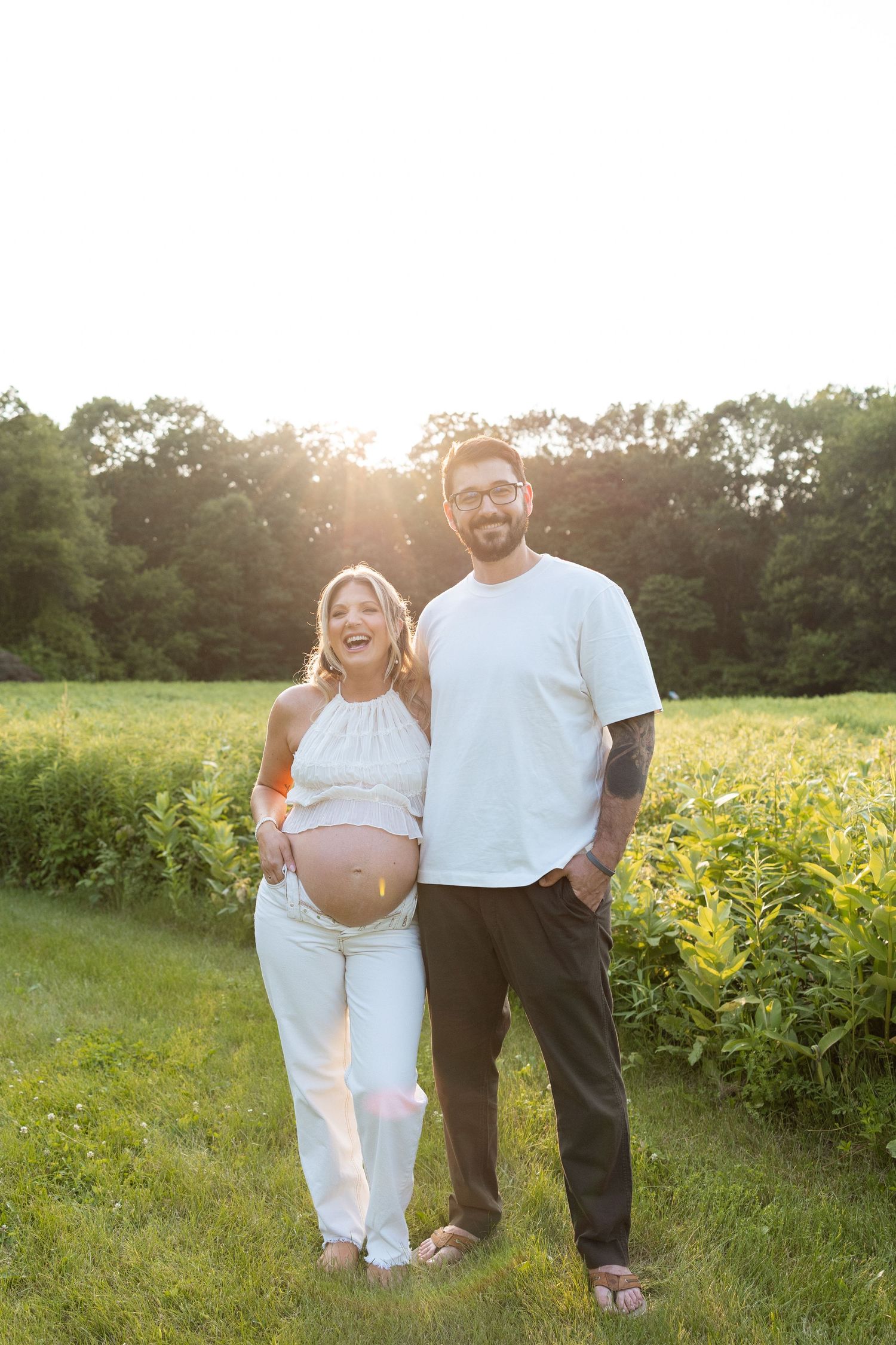 Expectant couple poses in a sunlit field at golden hour, with lush greenery and trees in the background.