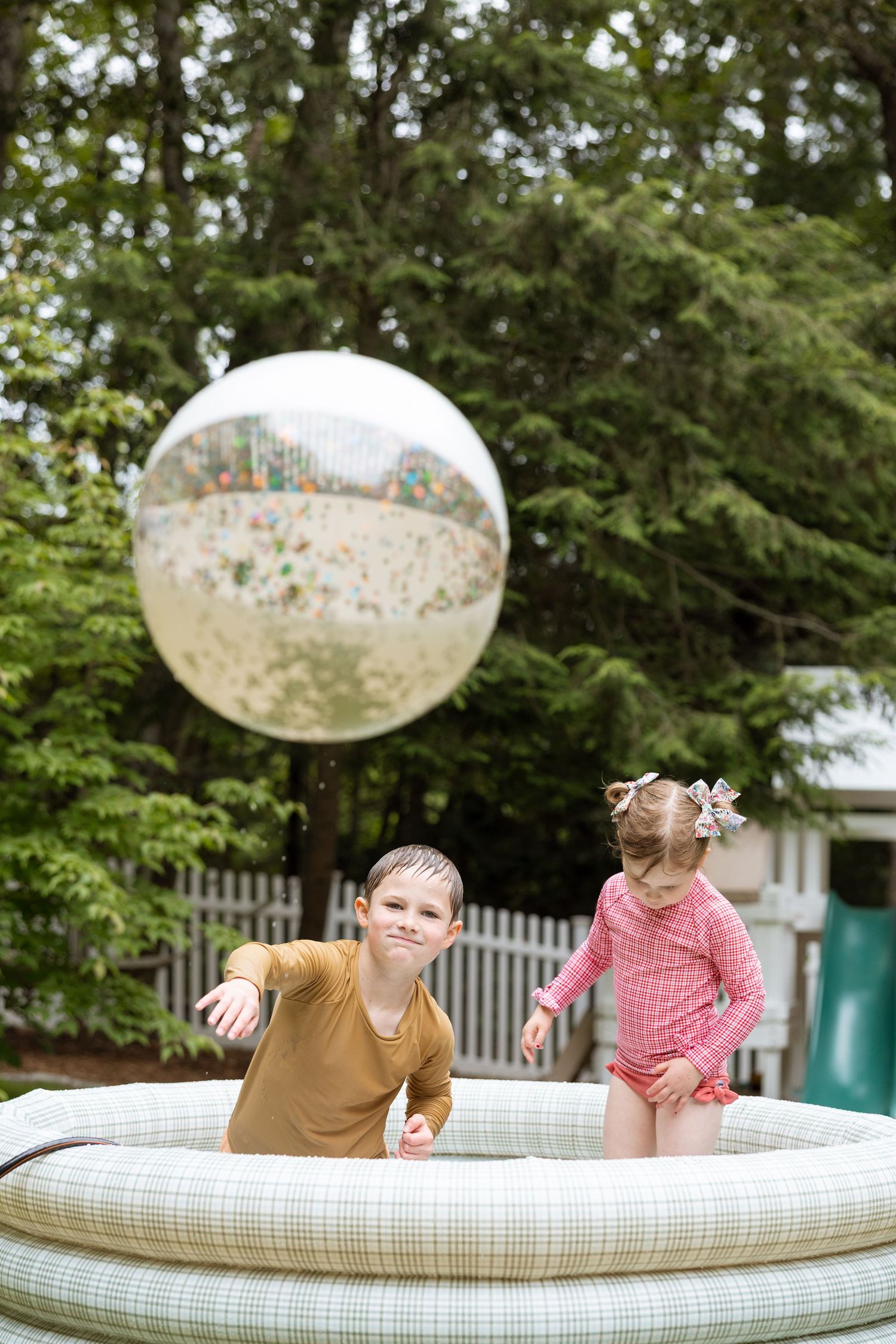 Two children play together on an inflatable bounce pillow cushion in a backyard garden setting.