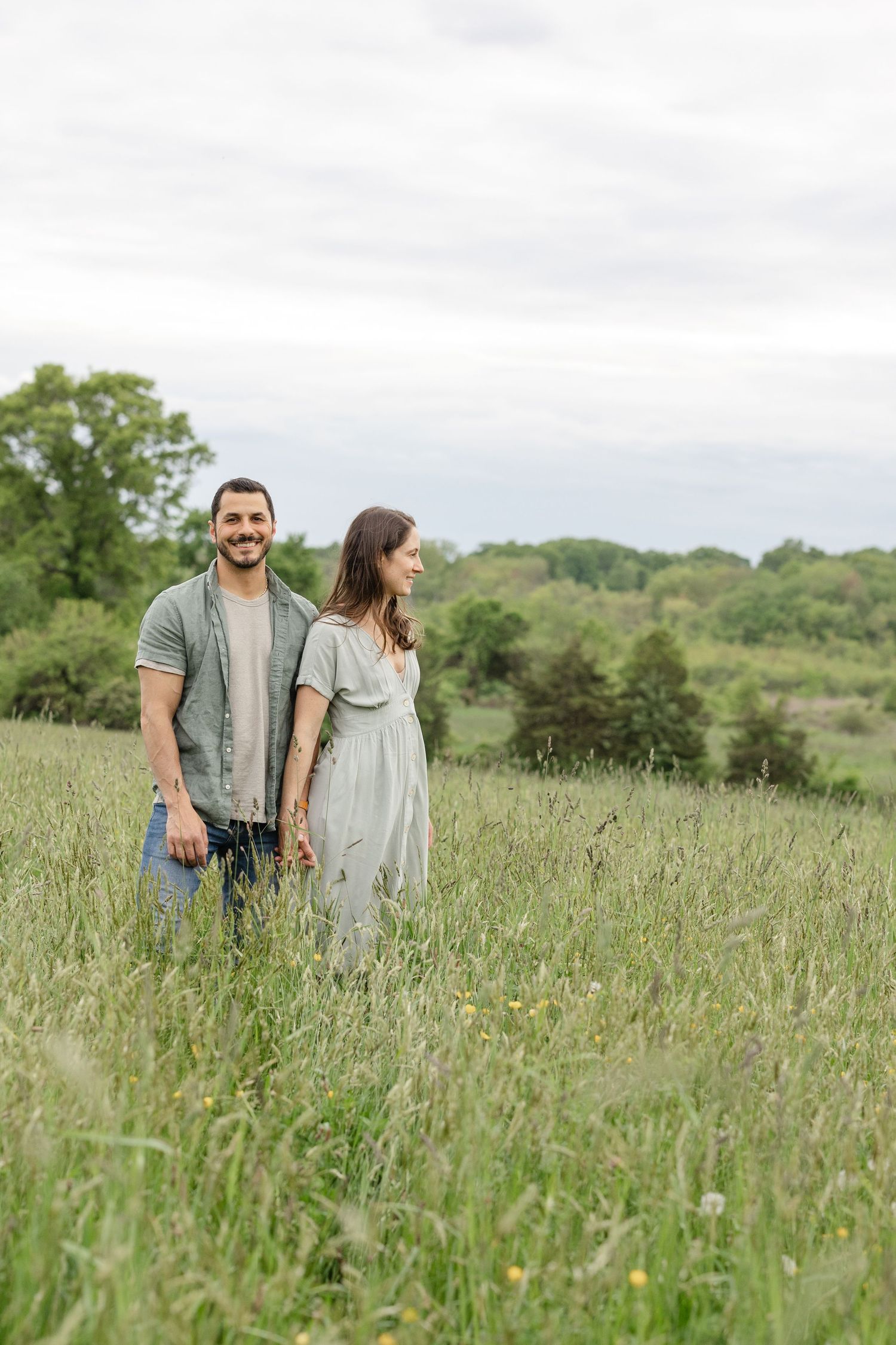 A couple stands together in a serene wildflower meadow surrounded by rolling green hills on a sunny day.
