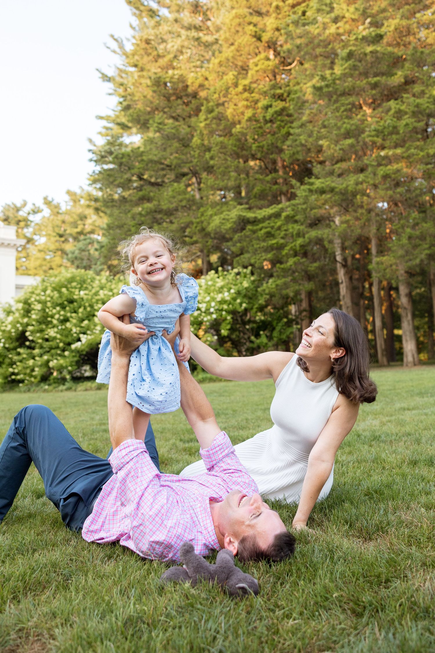 A joyful family plays together on grass with trees in the background on a sunny day.