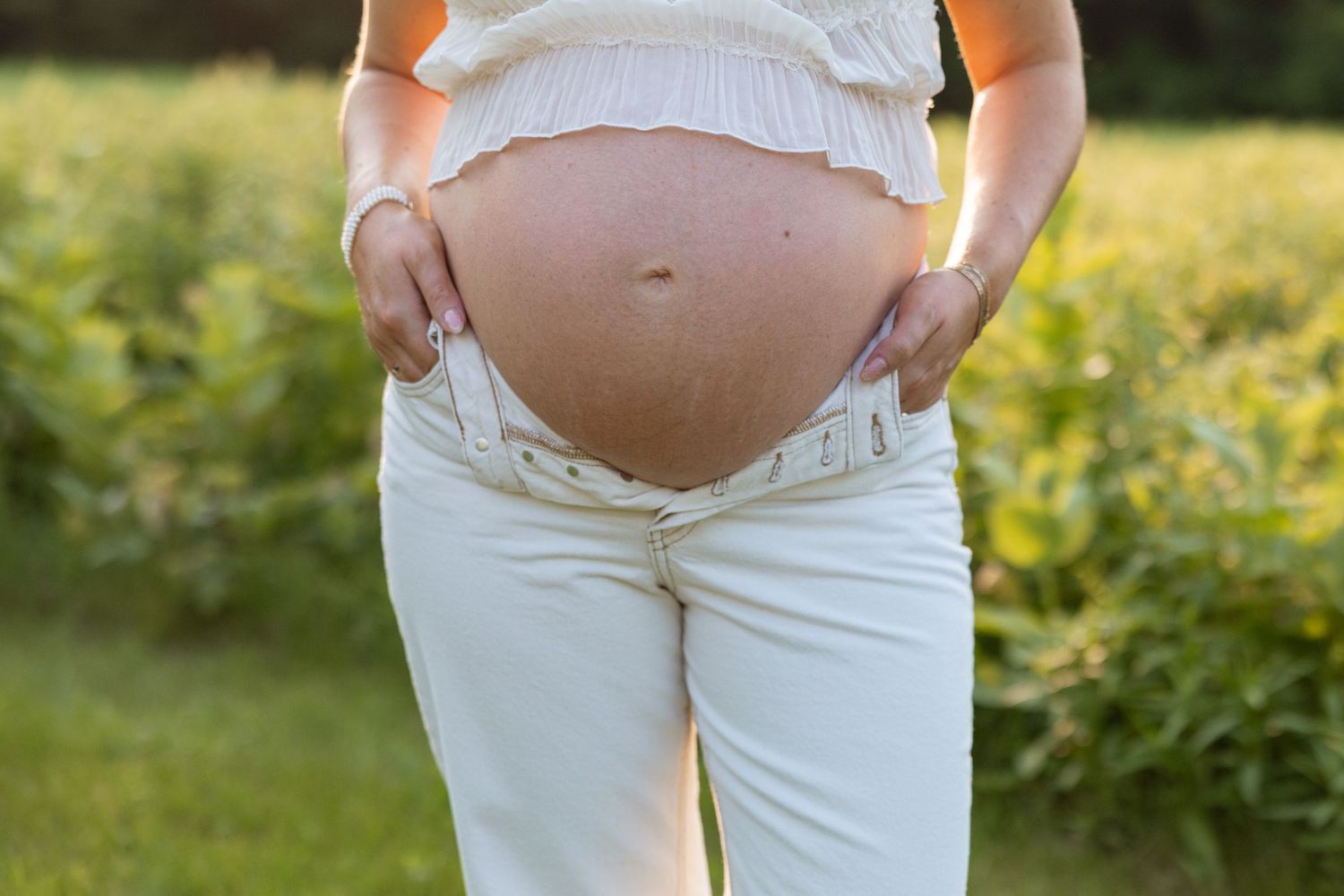 Close up of a pregnant belly wearing white jeans and ruffled top while standing in a sunny garden with yellow flowers.