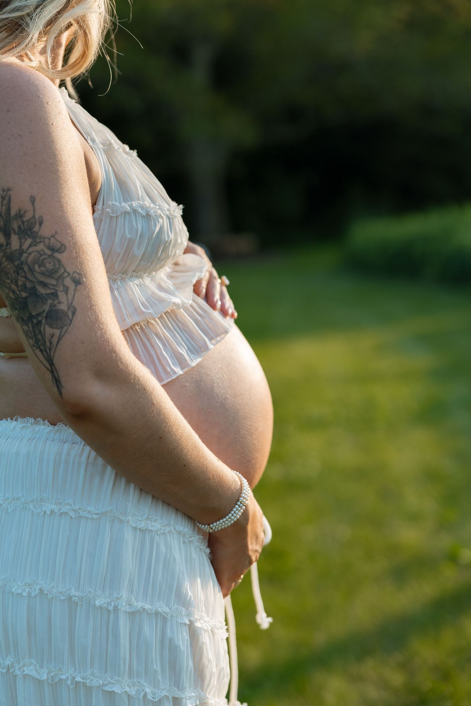 Side view of an expectant mother in a white lace dress with visible tattoos standing in a sunlit garden setting.
