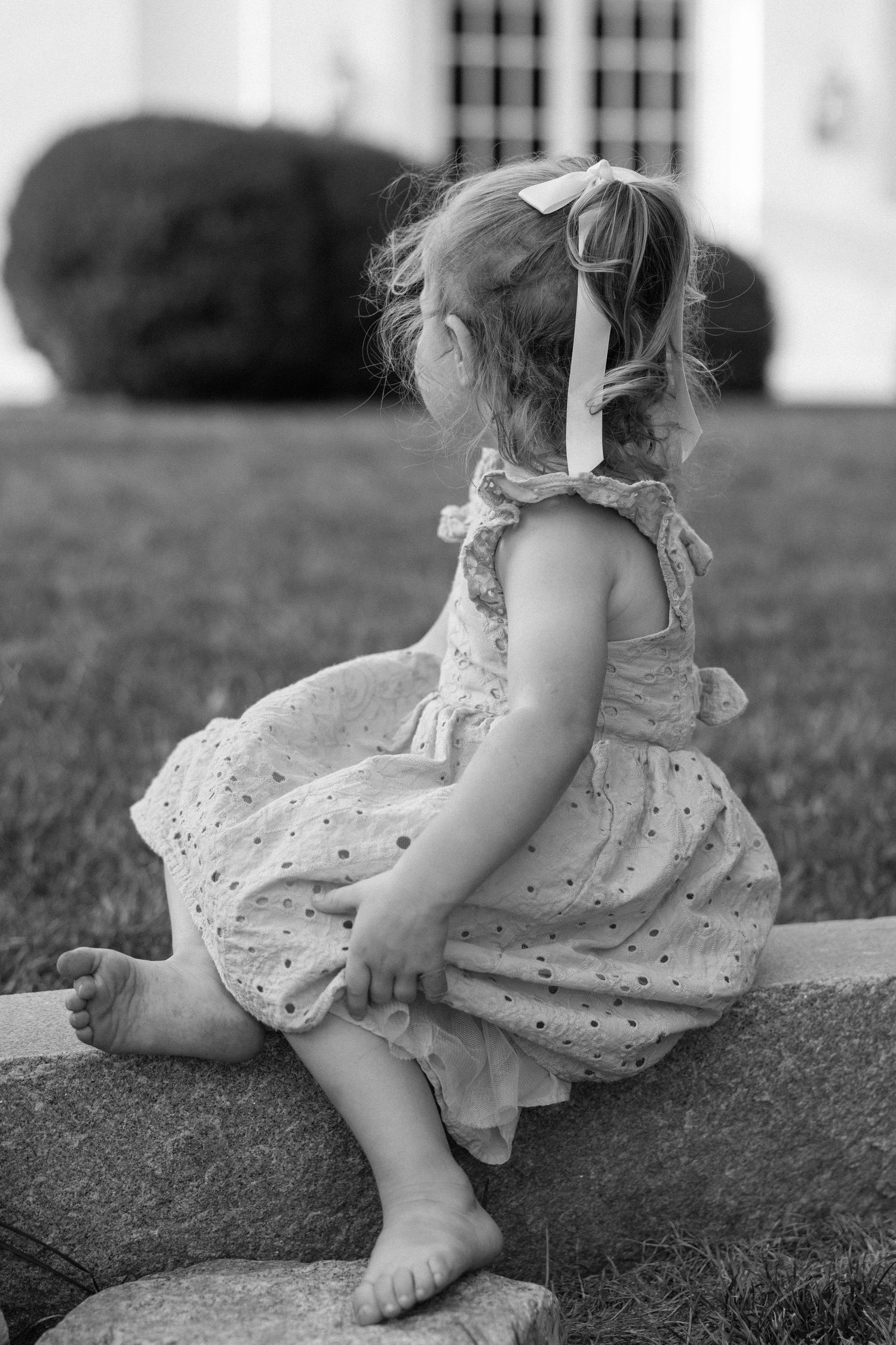A young child in a flowing sundress sits barefoot on a rock in black and white photography.