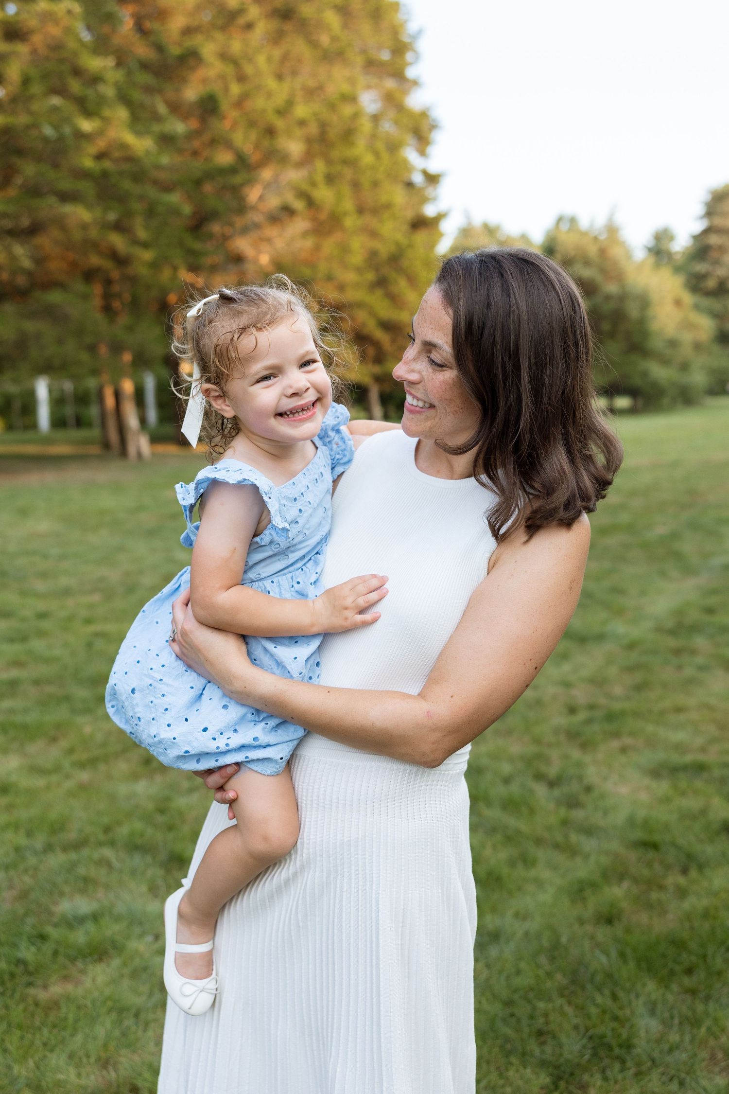A laughing child in a blue dress being held by someone wearing white in an outdoor park setting during summer.