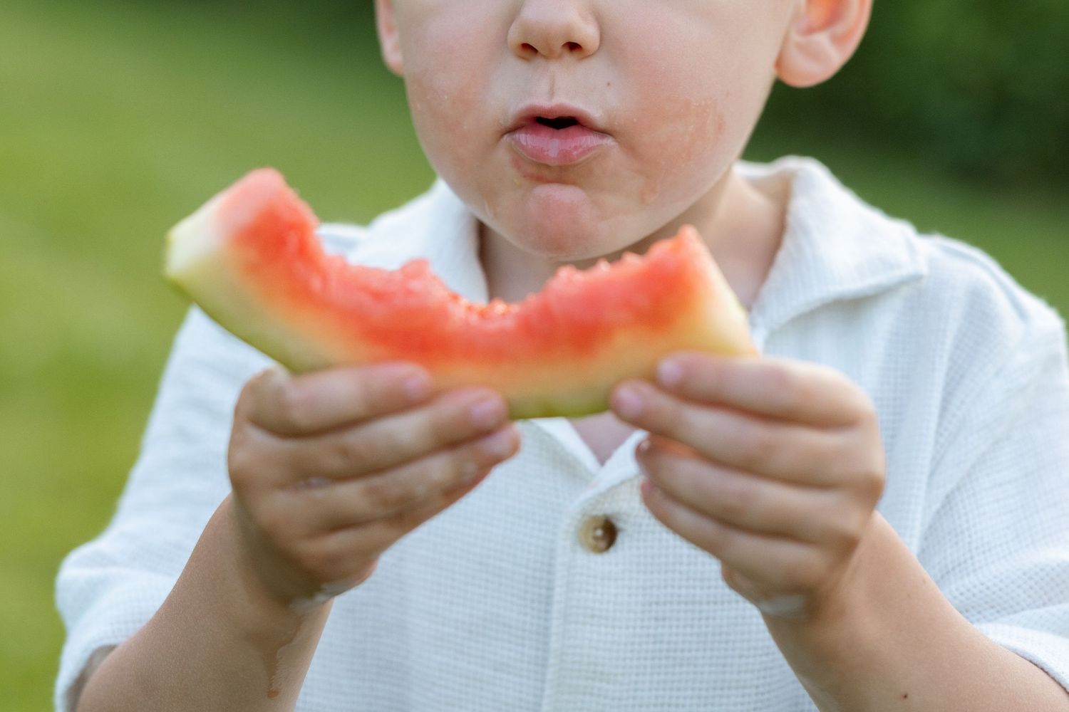 A close-up view of someone eating a fresh slice of watermelon while wearing a white shirt outdoors.