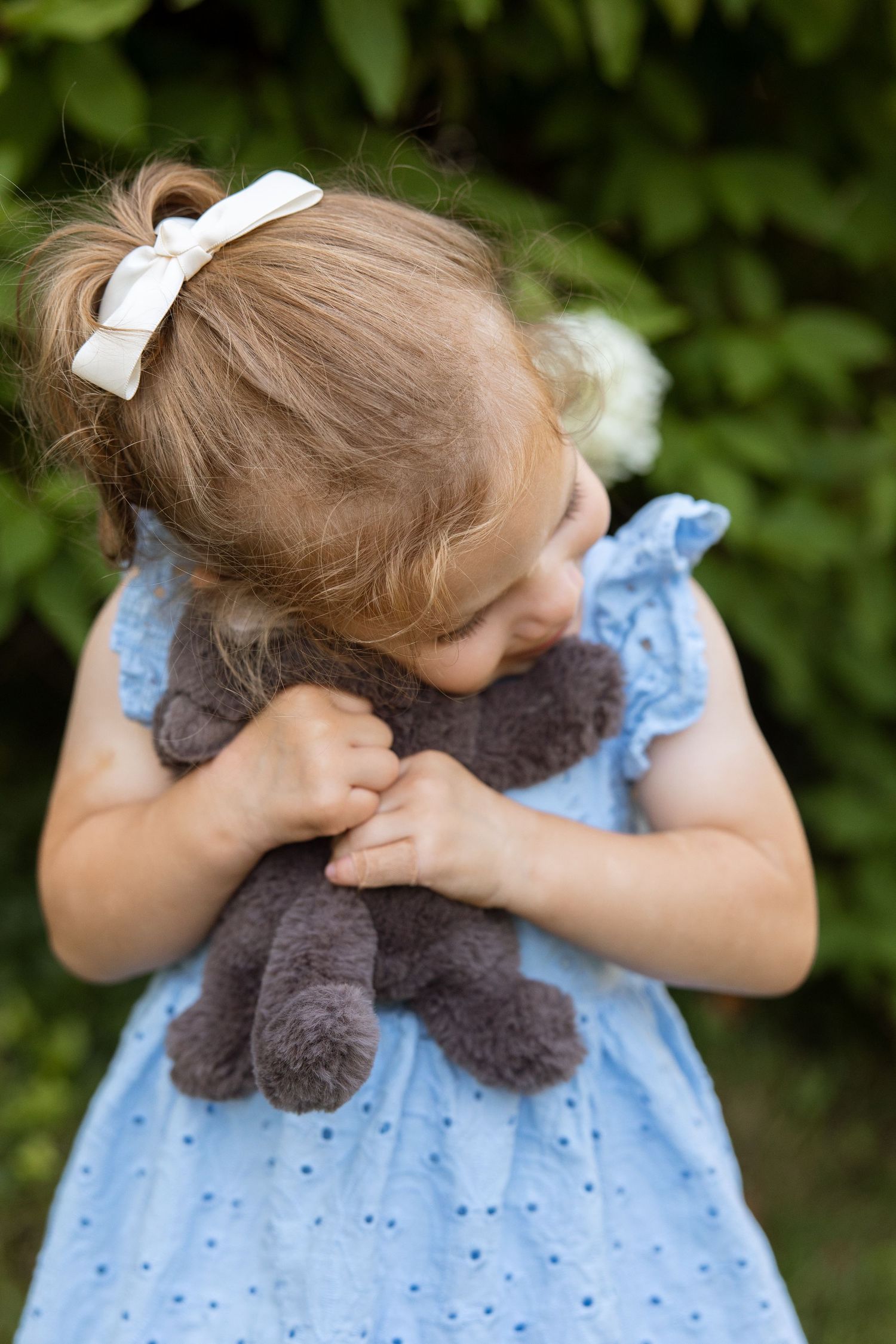 Young child in blue dress hugs gray teddy bear while wearing white bow in hair outdoors.