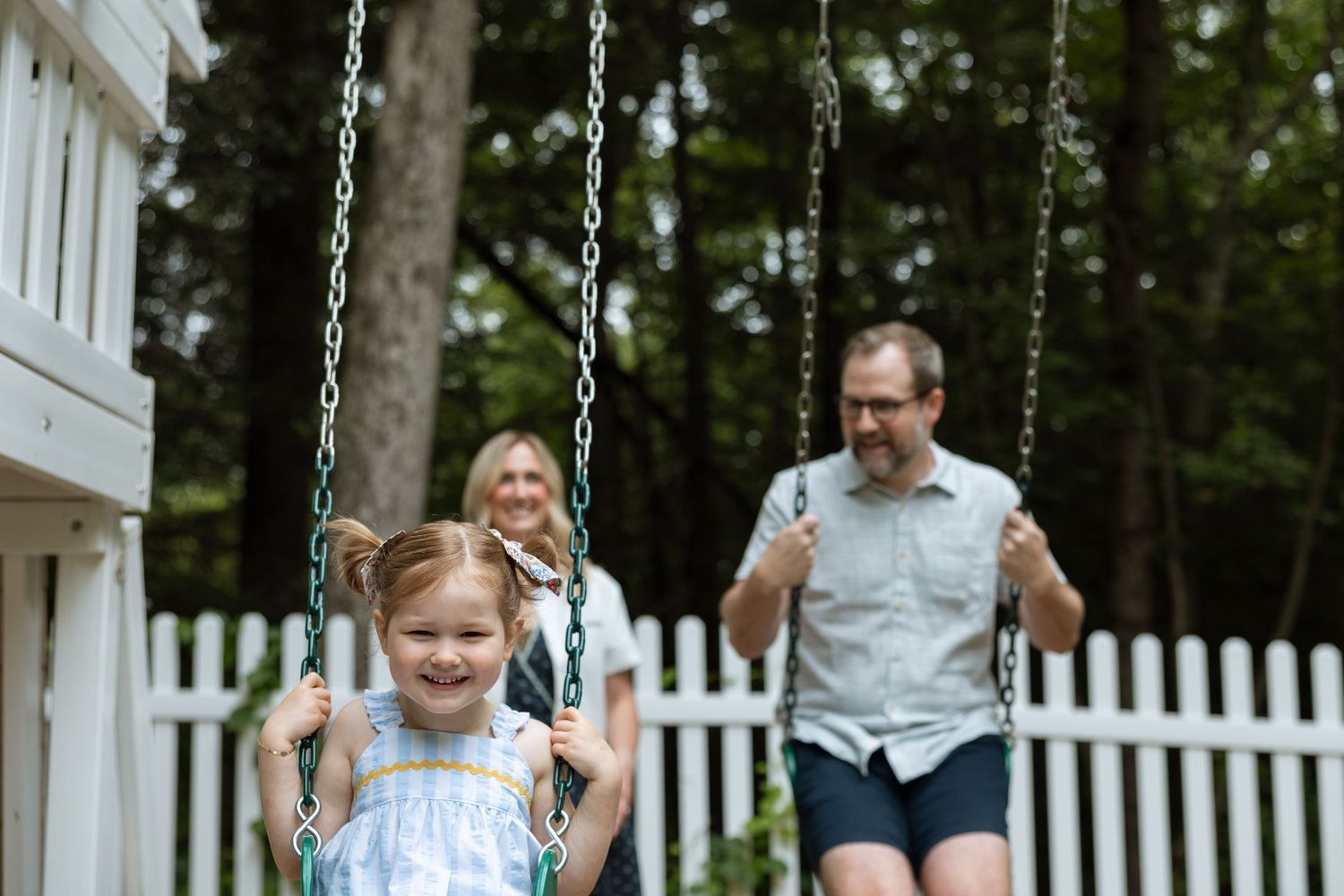 Children swing together on playground swings while adults supervise in a fenced backyard area.