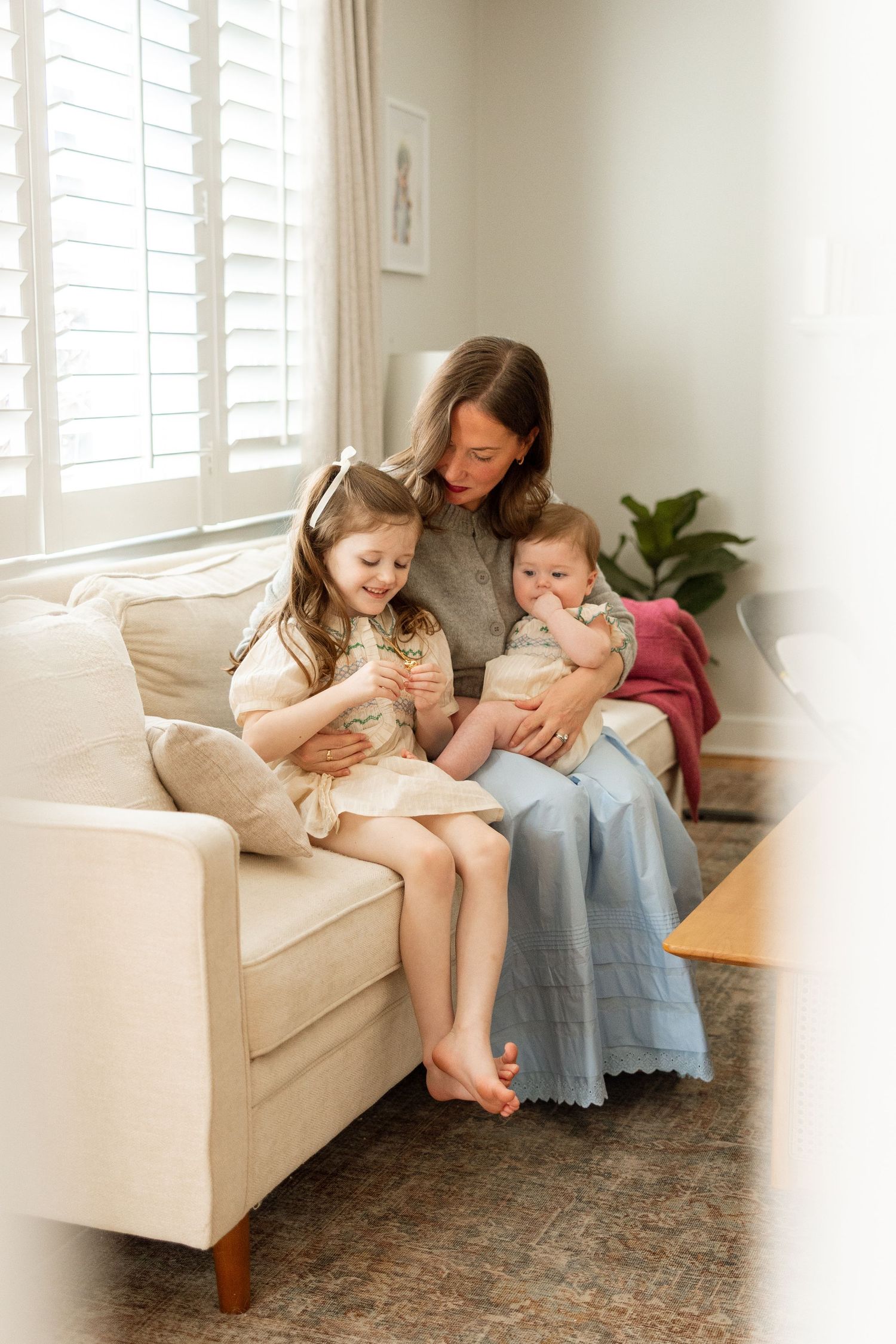 Three children sit together reading books on a cream-colored couch in a bright, sunlit room with white window shutters.