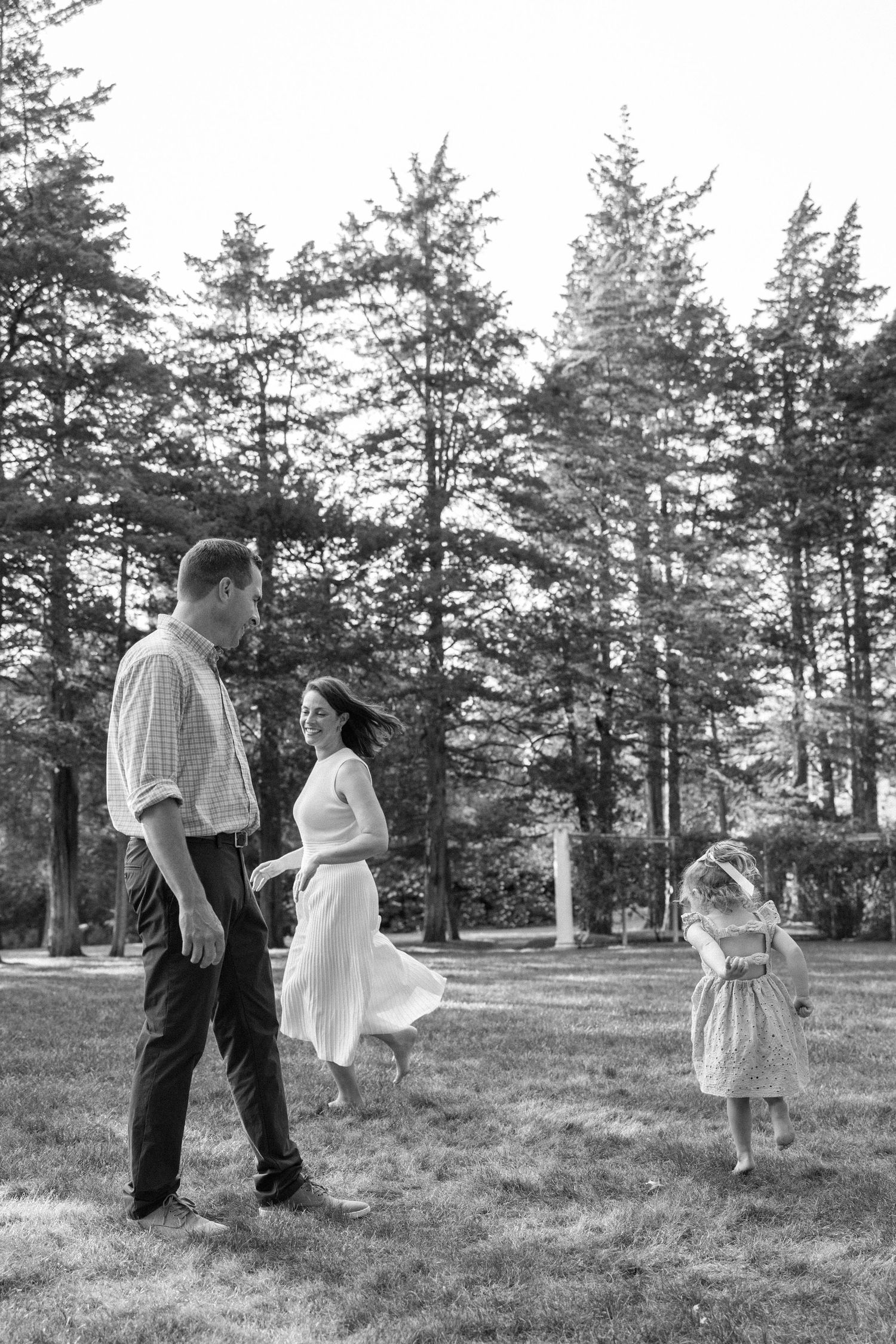 A family enjoys a candid moment together on a grassy lawn surrounded by tall evergreen trees in black and white.