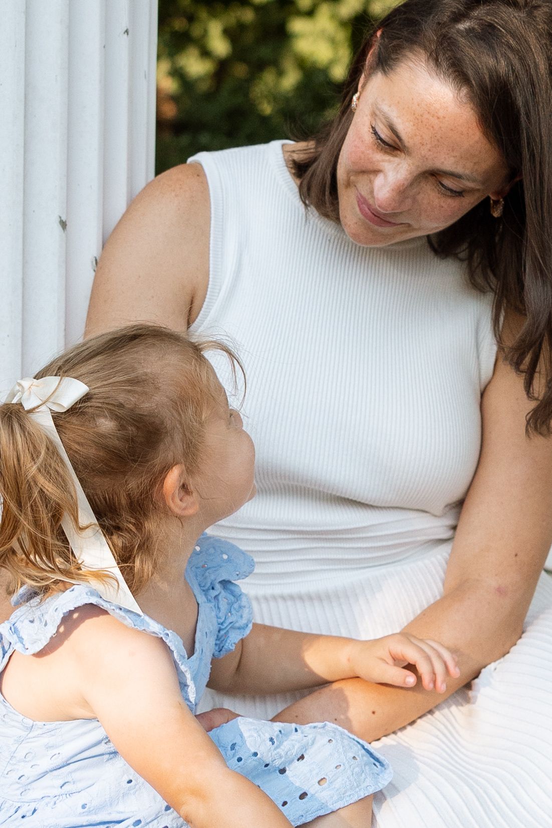 A tender moment between an adult in white and small child in blue dress sitting together outdoors.