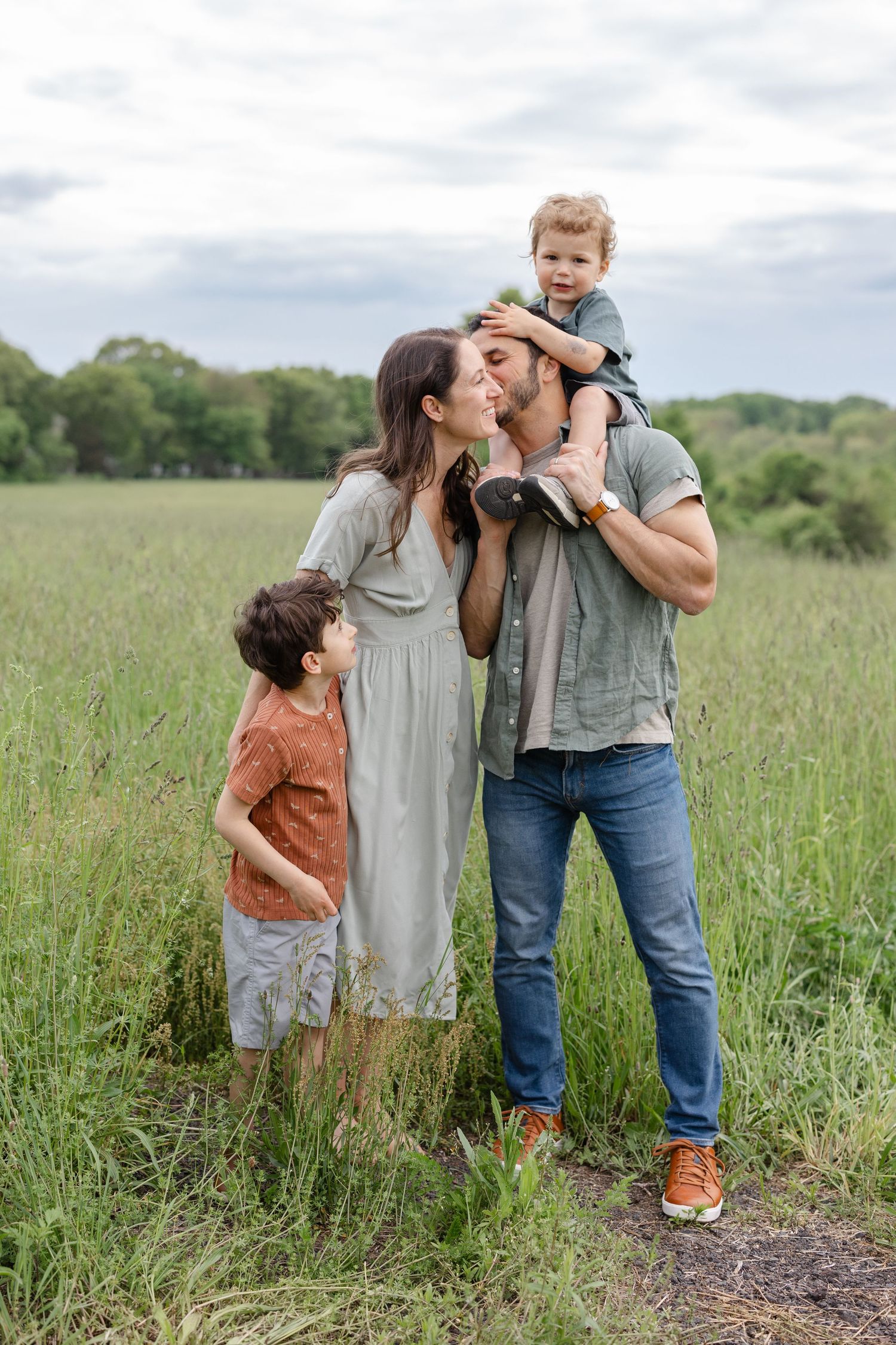 A family enjoys a tender moment together while standing in a tall grass field on a cloudy day.