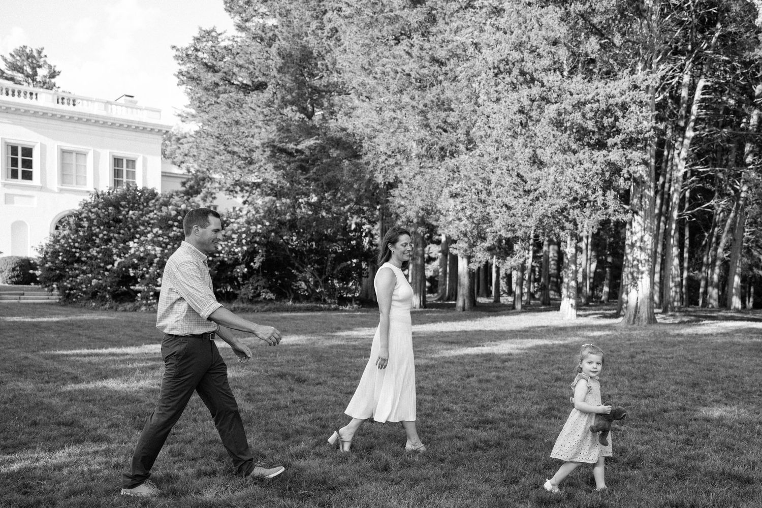 A black and white photo of a family walking together across a lawn near trees and a house on a sunny day.