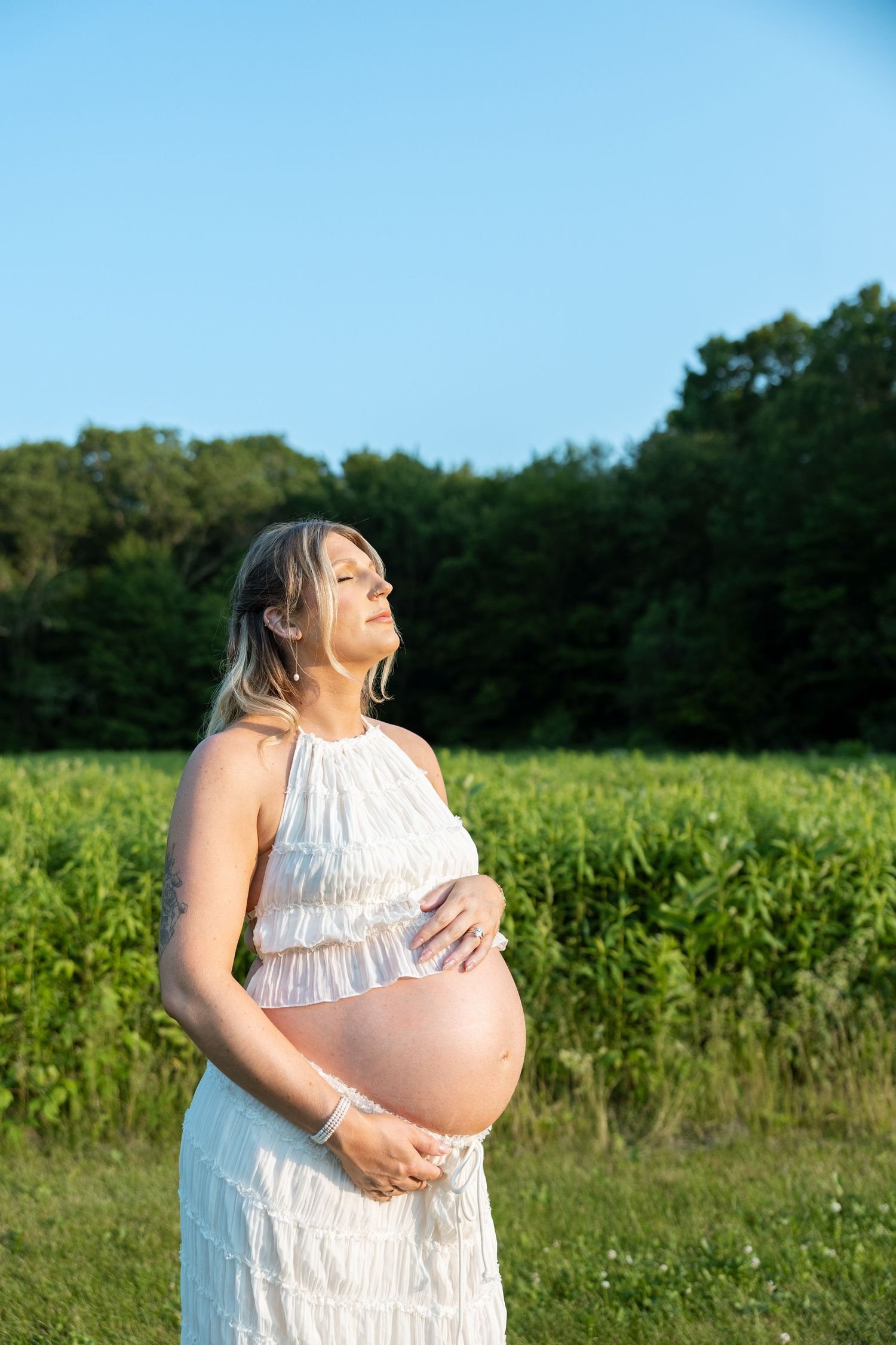 Woman in white ruffle dress poses for maternity photos in a sunlit field near a cornfield.