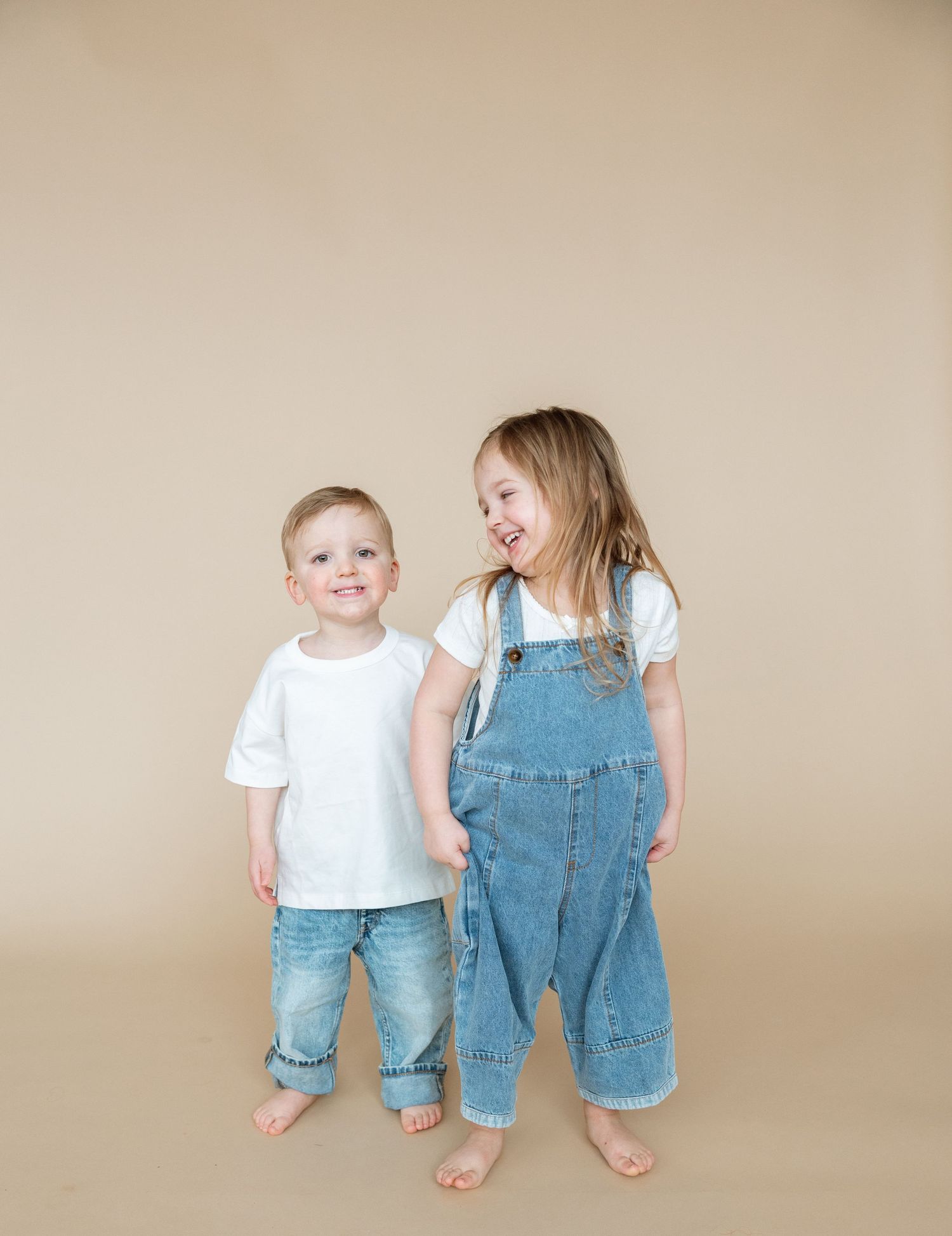 Baby in white shirt and denim overalls sits next to toddler in matching outfit against neutral background.