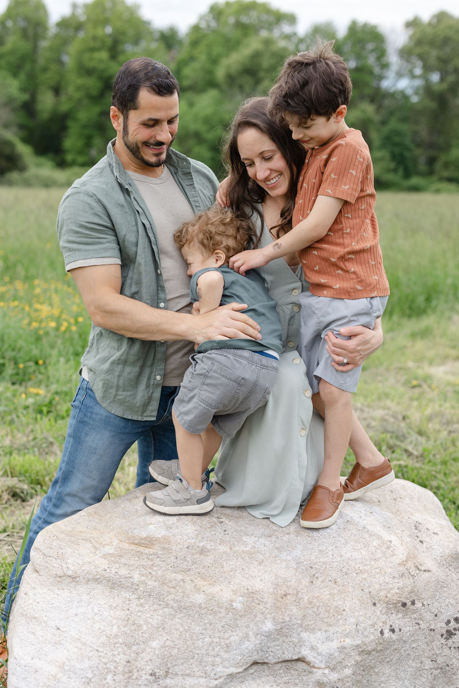 Family wearing casual clothes stands together on large boulder in sunny meadow with green trees in background.
