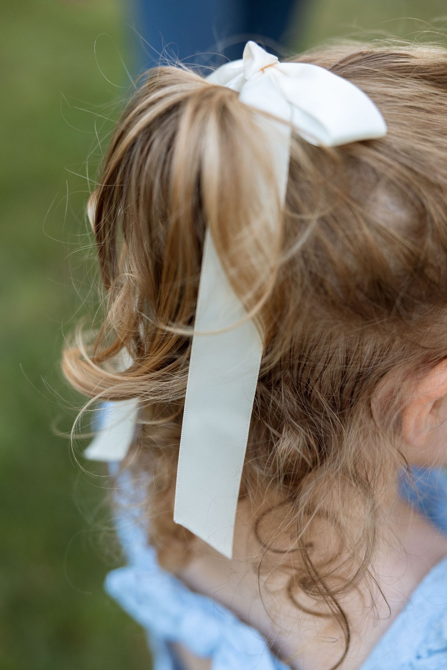 Light brown curly hair adorned with a white ribbon bow and blue fabric dress visible in profile view.