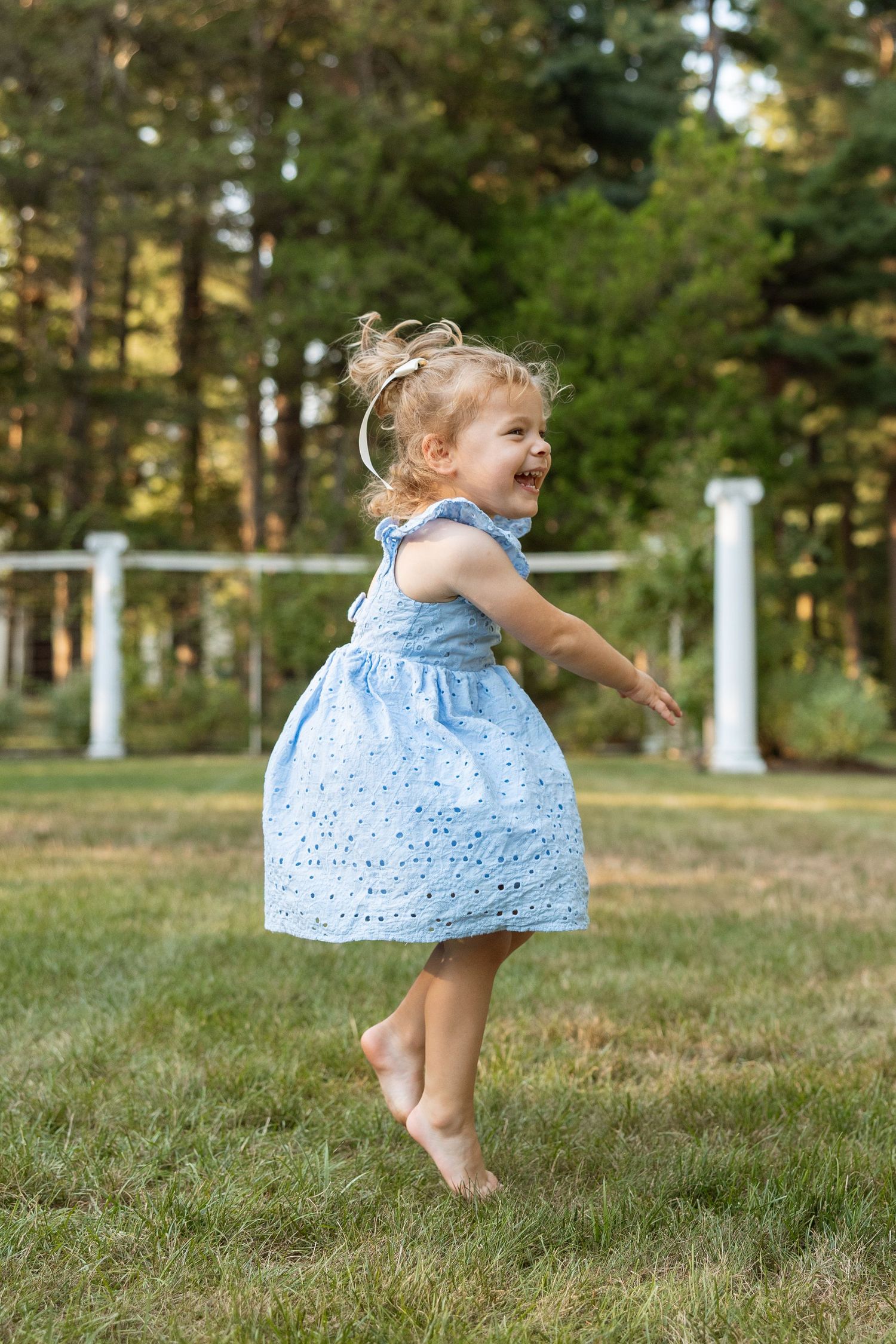 Small child in light blue dress twirls joyfully on grass during a sunny summer day in a garden setting.