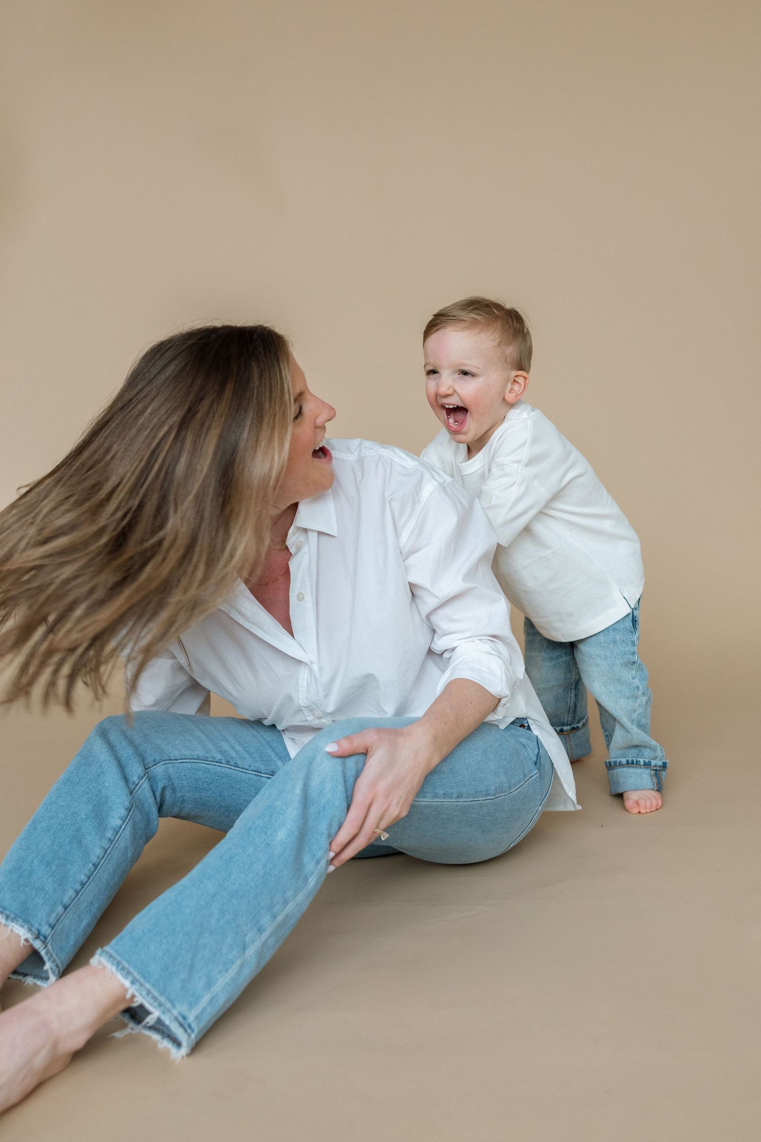A playful moment between parent and child sitting on a beige background wearing matching white shirts and jeans.