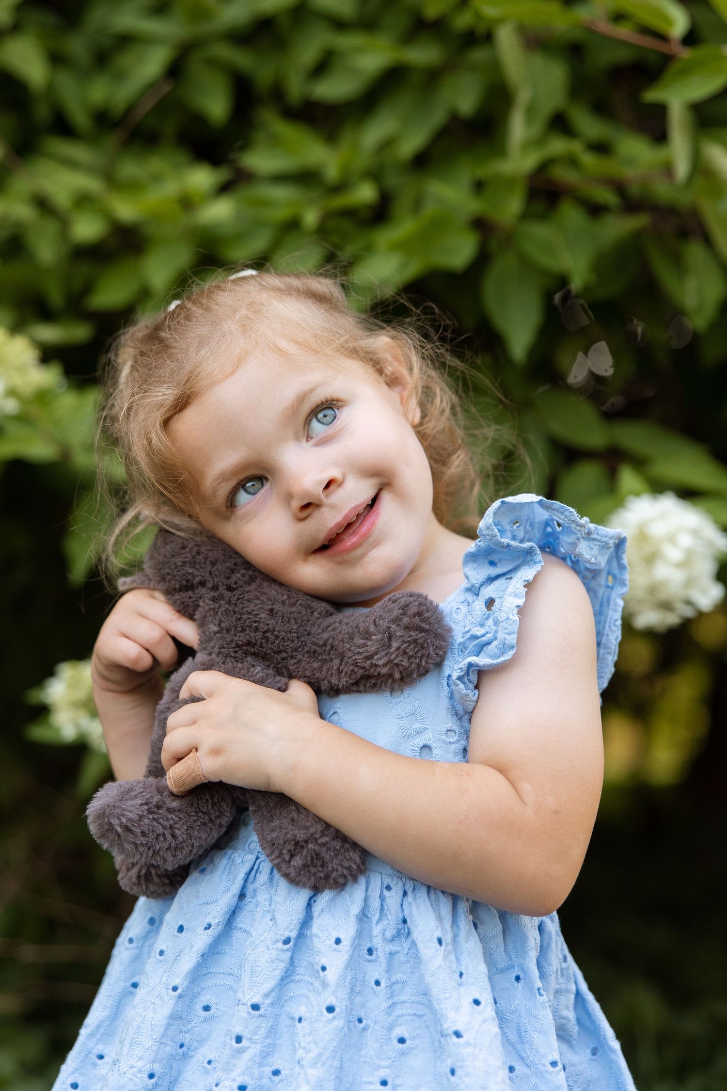 Happy child in blue polka dot dress hugs stuffed animal toy outdoors with green foliage background.