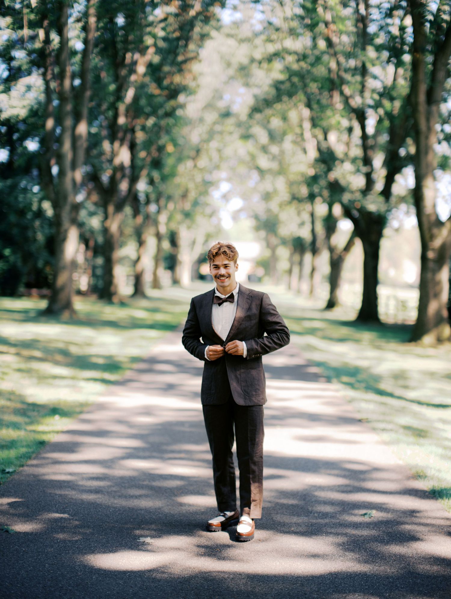 Person in formal attire standing on a tree-lined path in dappled sunlight.