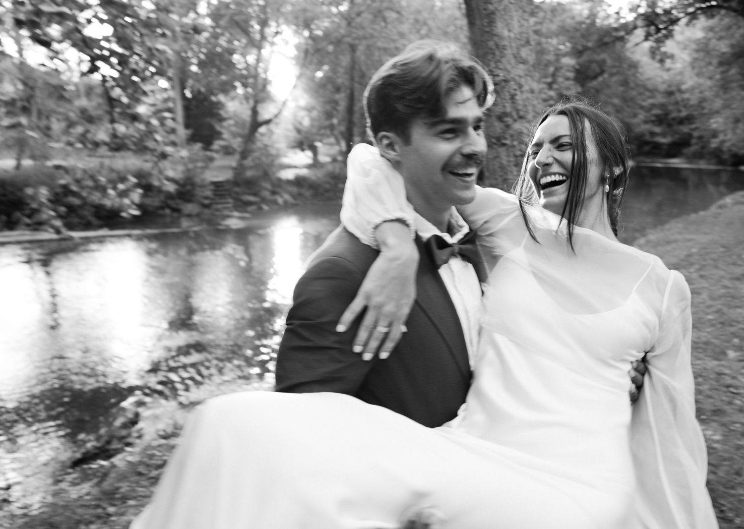 Joyful wedding couple laughing by a pond with bride being carried.