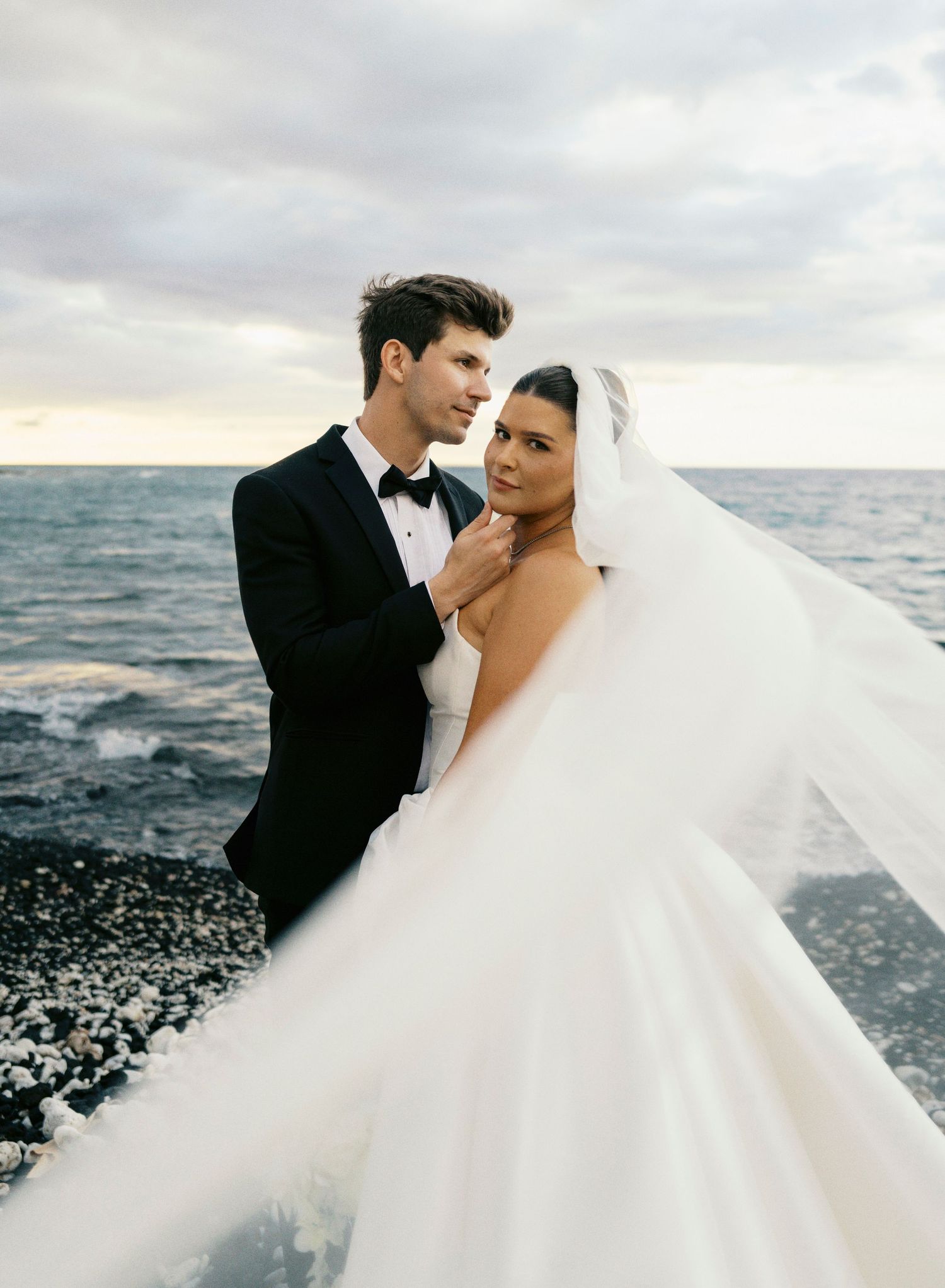A romantic wedding couple embraces on a rocky beach at sunset with a flowing white veil in the wind.