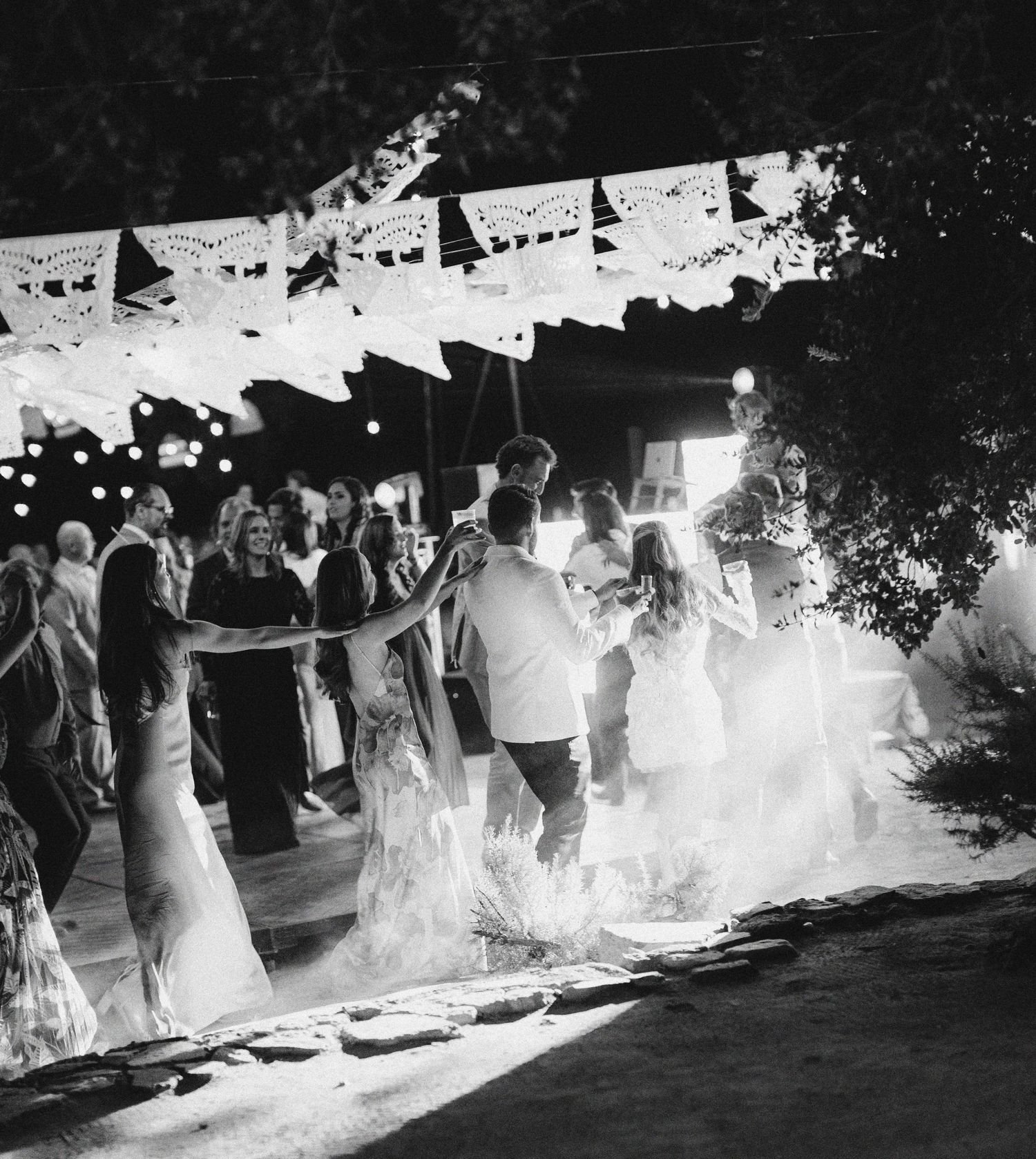 A group of wedding guests dance under string lights at an outdoor evening reception as smoke fills the dance floor.