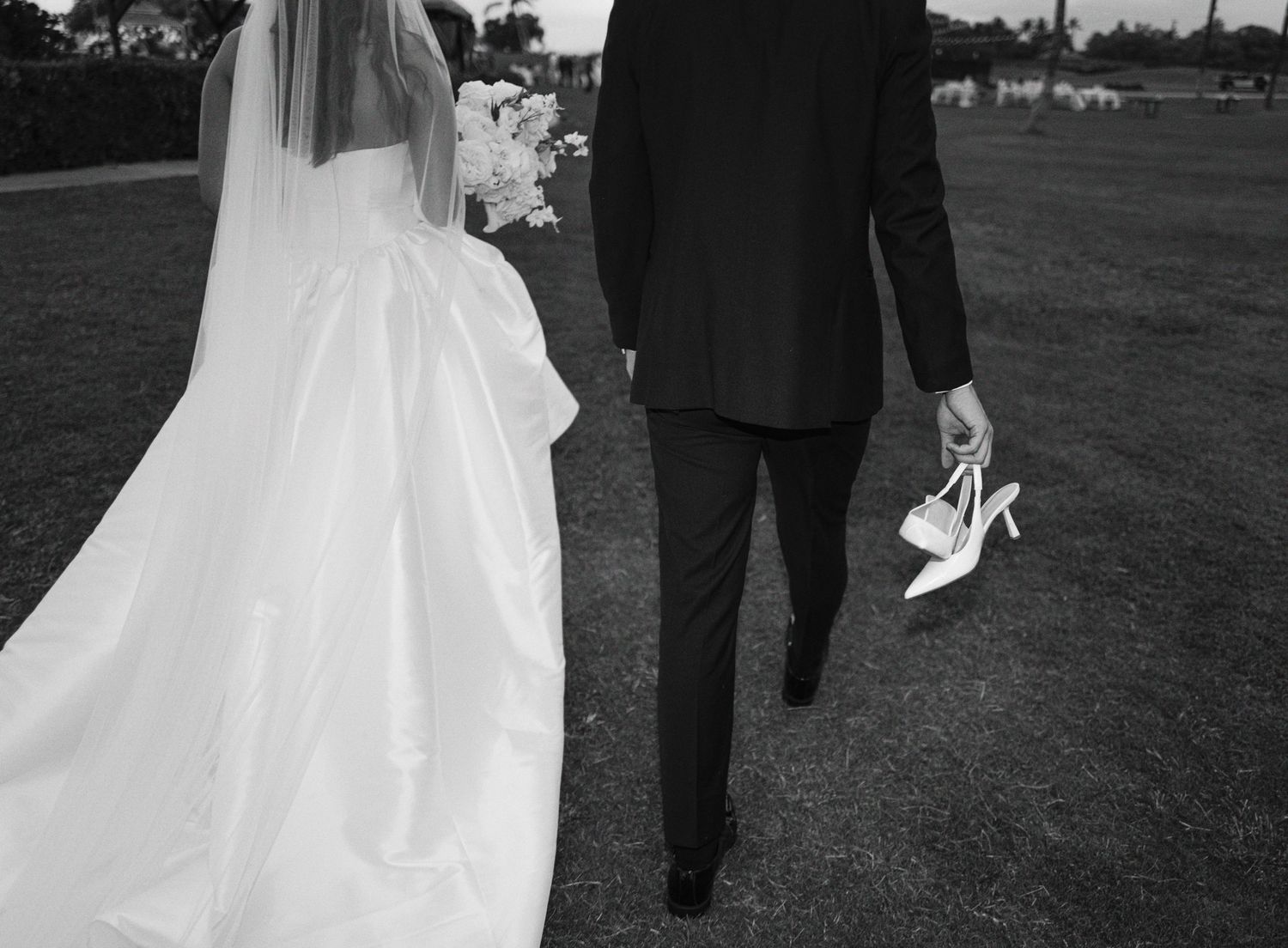 Wedding couple walks across grass lawn holding hands with flowing white bridal gown trailing behind them.