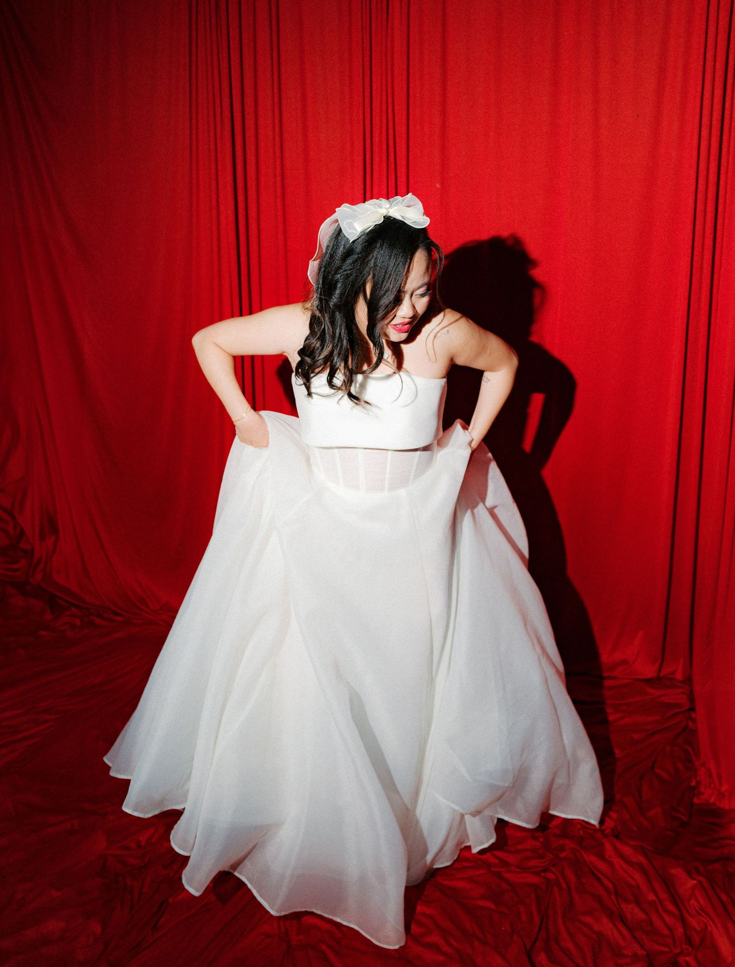Bride in flowing white gown twirls against red backdrop creating dramatic shadows.