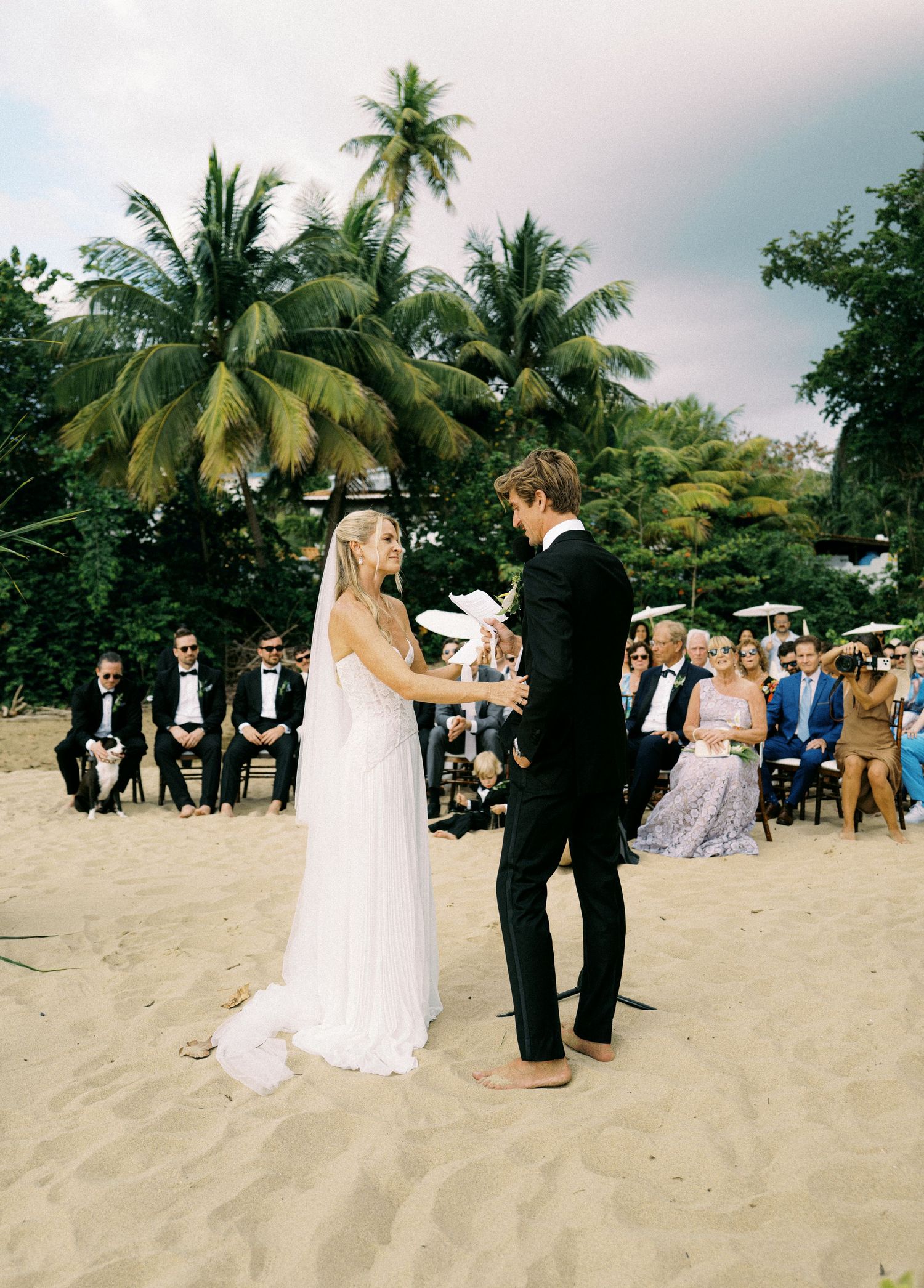 A beach wedding ceremony takes place under palm trees with guests gathered on the sandy shore at sunset.
