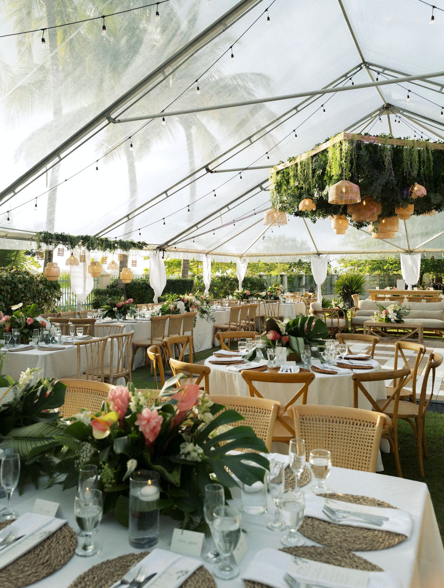 Elegant wedding reception tent setup with wooden chairs, hanging lanterns, and tropical greenery decor at sunset.