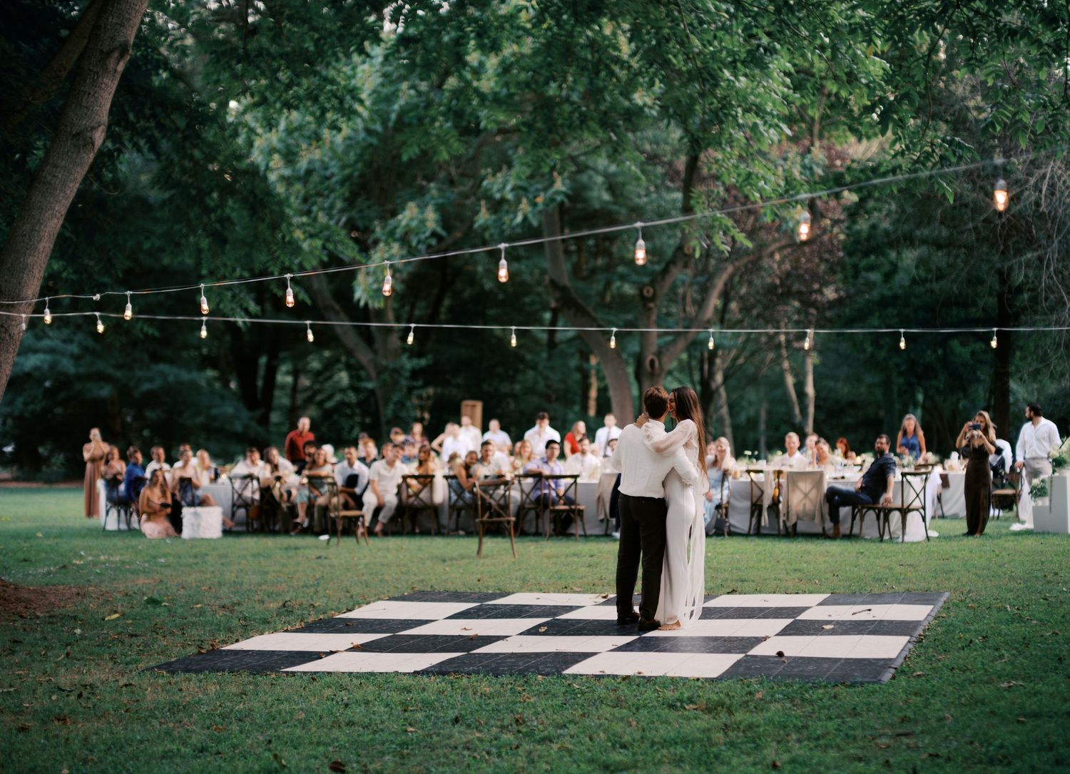 Outdoor reception area with string lights and white dining tables arranged under tall trees at dusk.