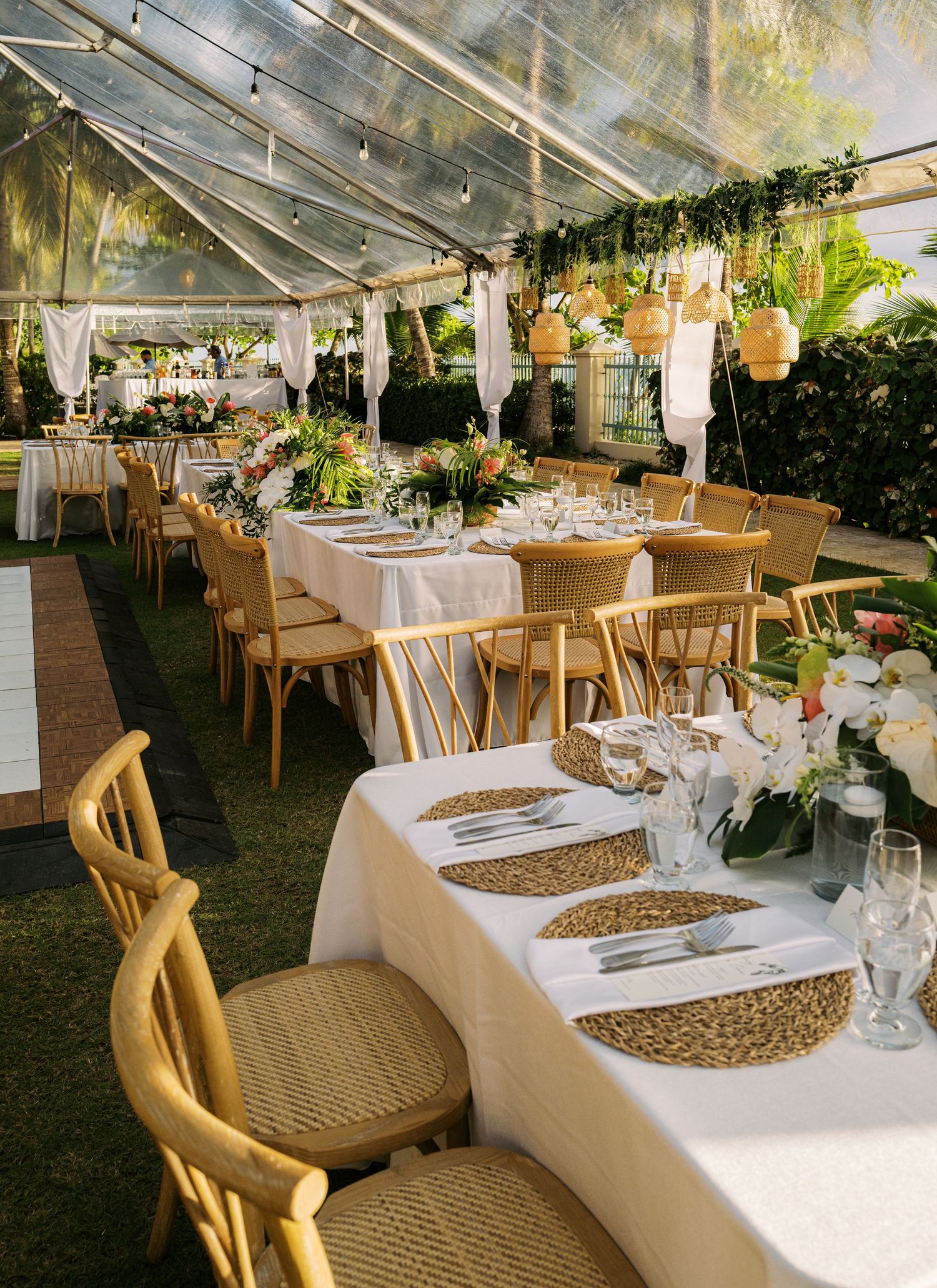 Elegant table settings with gold charger plates and floral centerpieces under a clear tent for an outdoor wedding reception.