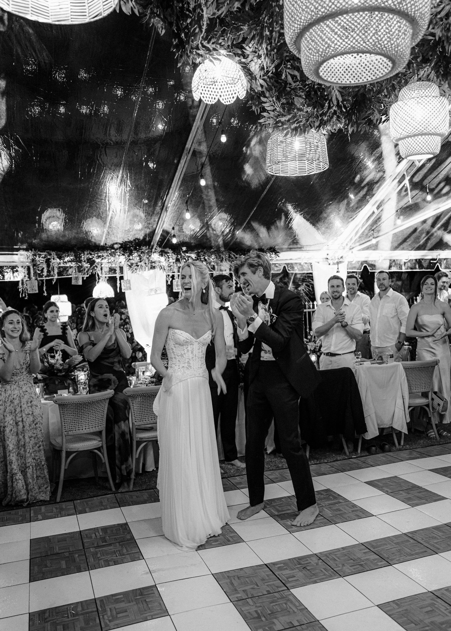 A wedding couple dances together on a checkered dance floor at an outdoor evening reception under hanging lights.