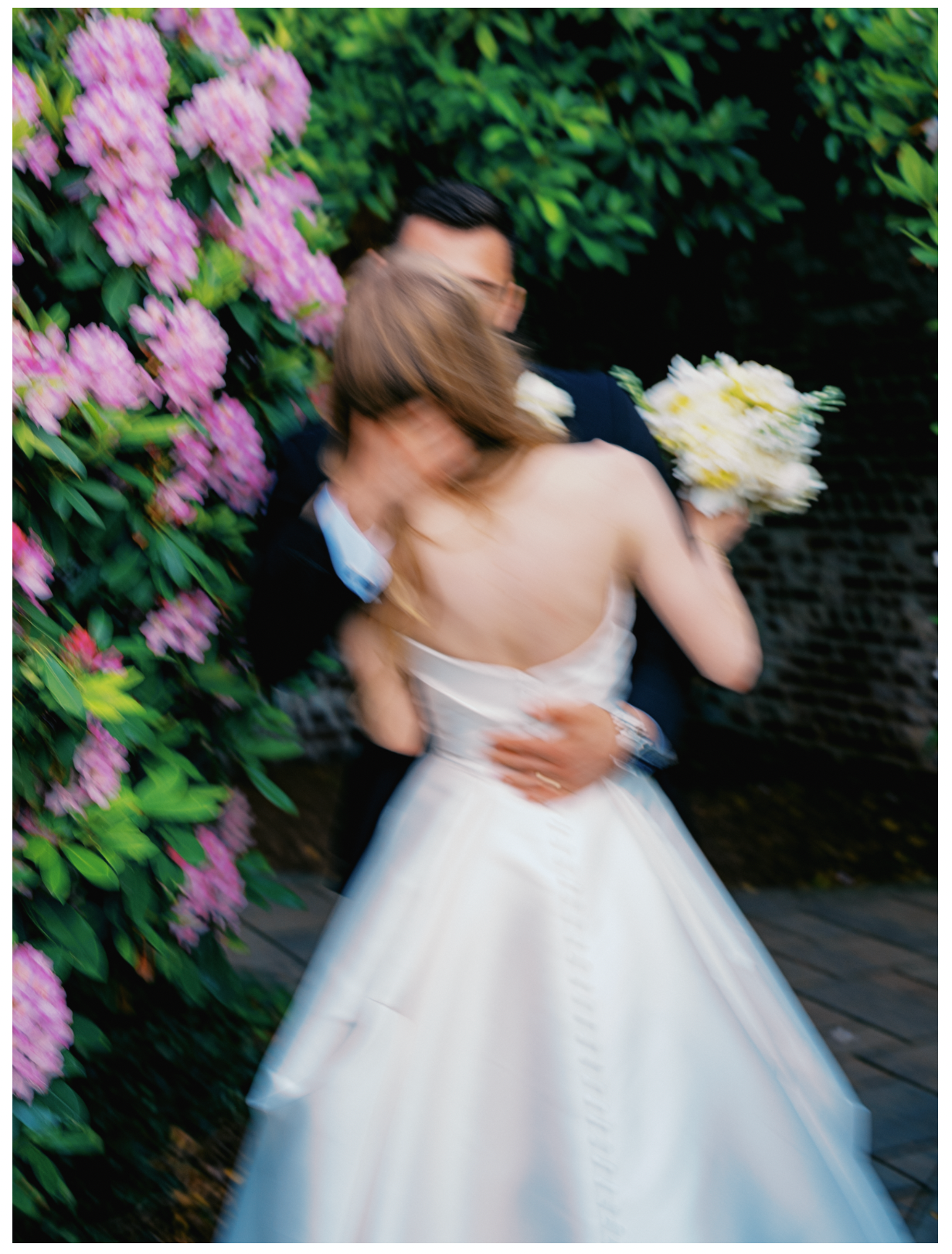 A bride in a white ballgown holds a bouquet while embracing a groom in a navy suit among blooming pink flowers.