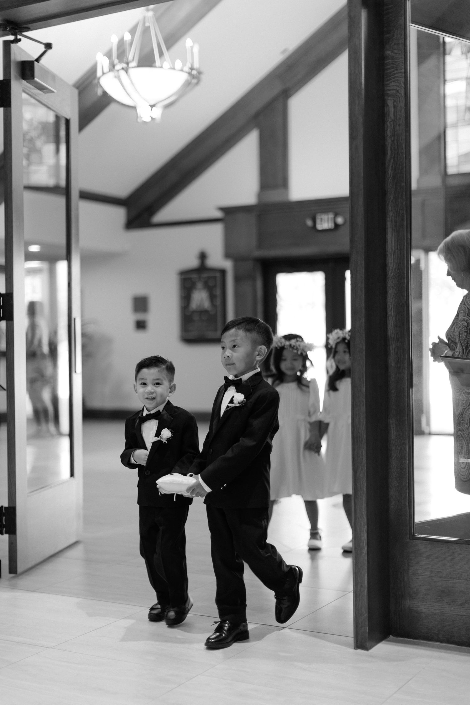 Black and white photo of young boys in tuxedos hanging playfully from beams in an indoor venue.