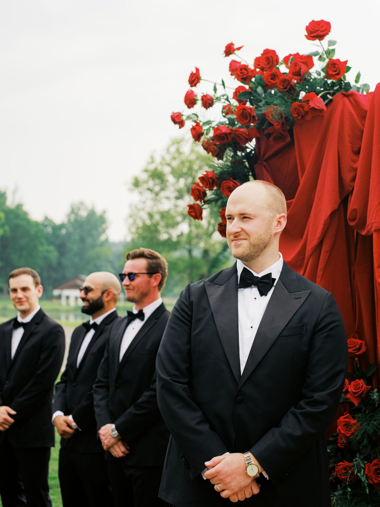 Groomsmen in black tuxedos stand outdoors with red rose decorations at an elegant wedding ceremony.