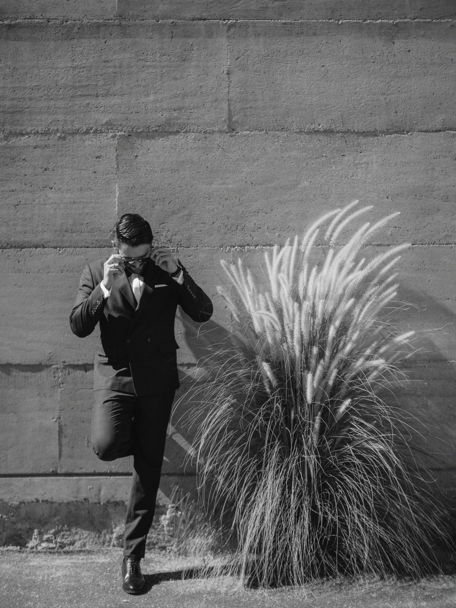 A photographer in dark clothing takes pictures of ornamental grass against a concrete wall in black and white.
