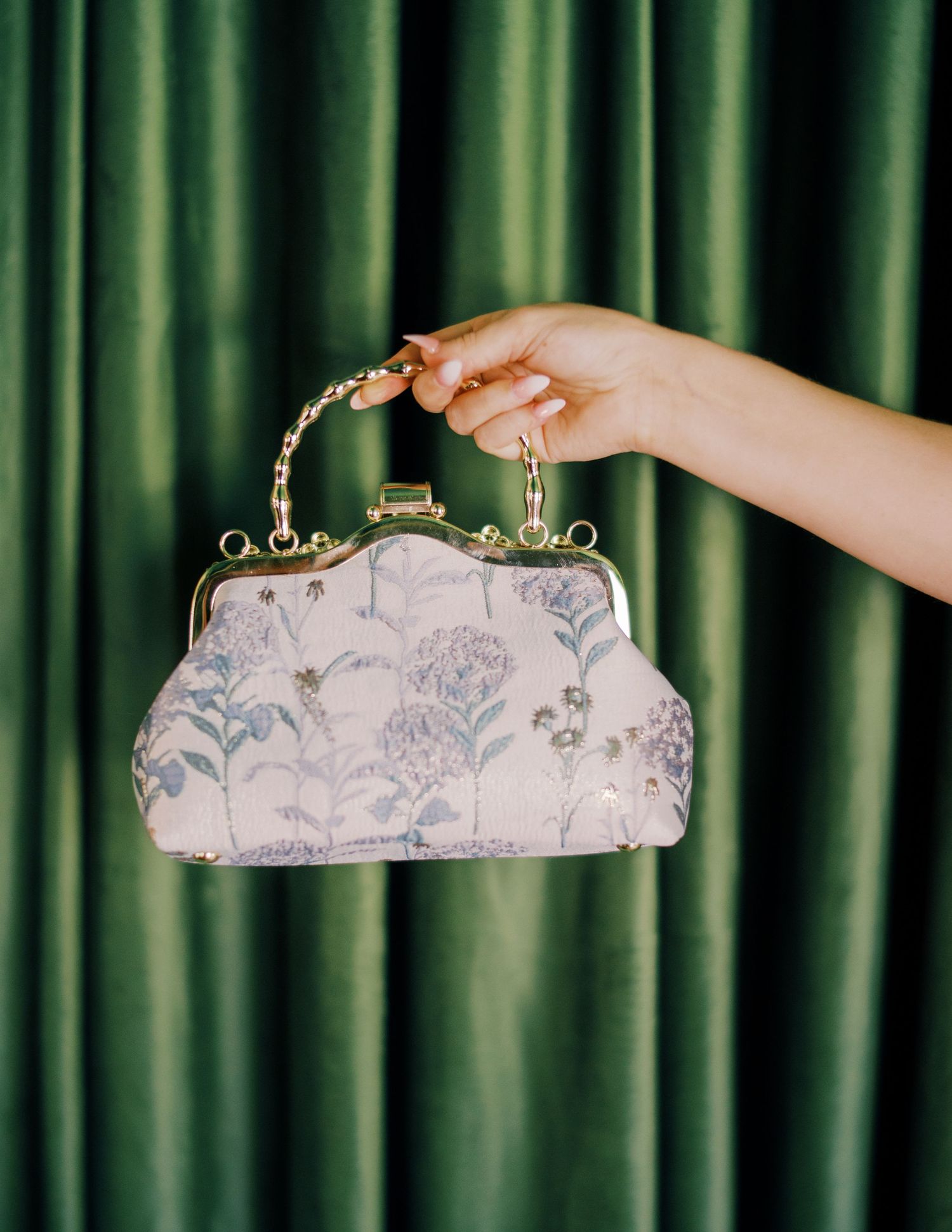 Three elegant pink floral clutch purses with metal clasps displayed in a row against a green curtain background.