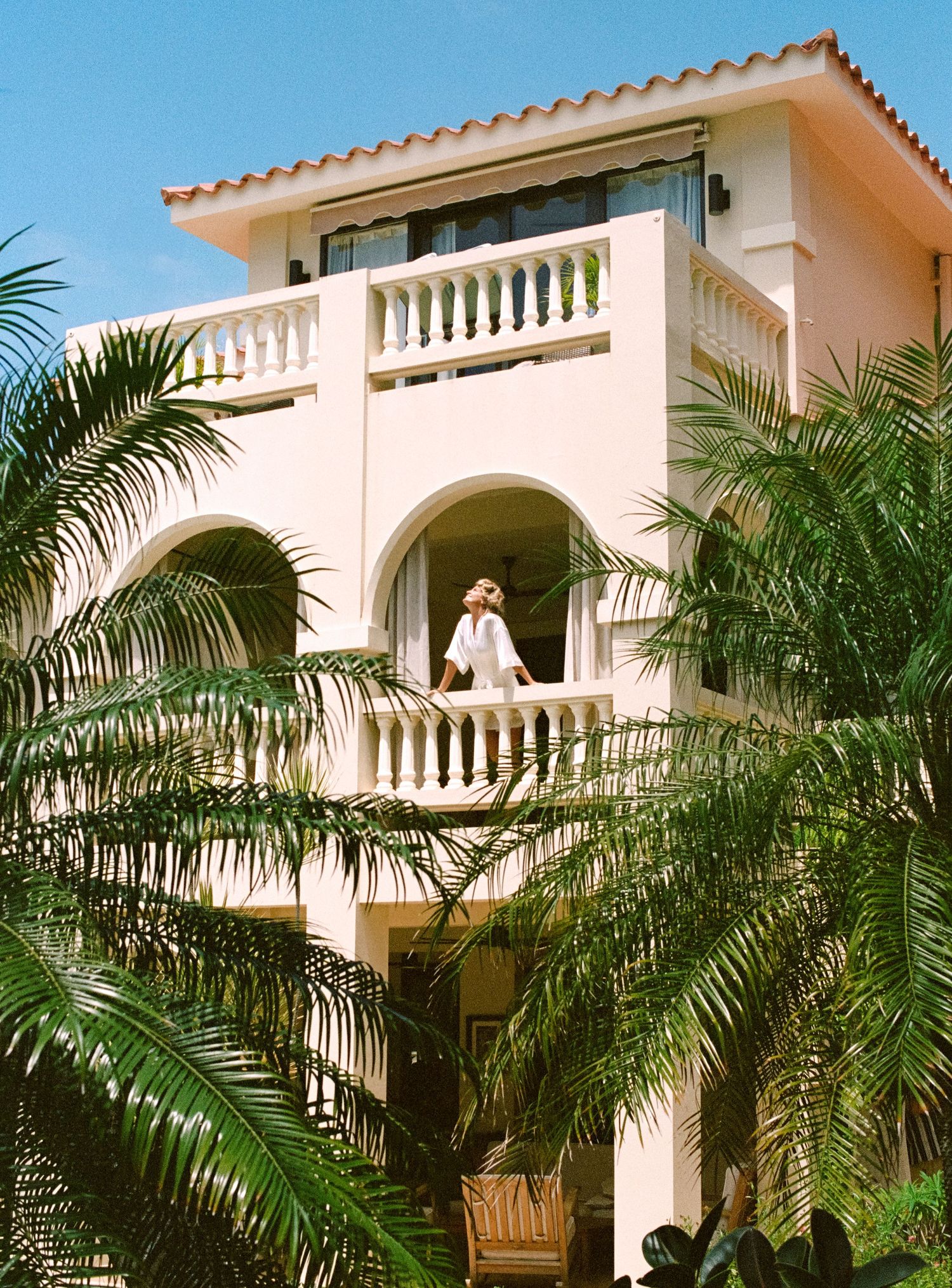 White Mediterranean-style villa with terracotta roof and lush palm trees framing the balconies in a tropical setting.