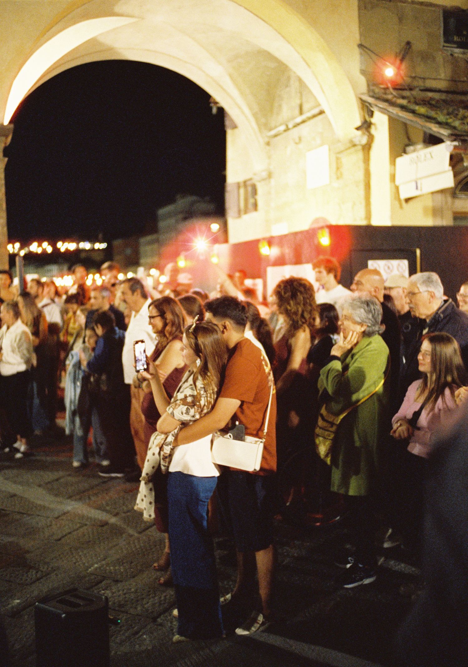 A crowd of people gathered under arched passageways at night with glowing red lights creating an atmospheric ambiance.