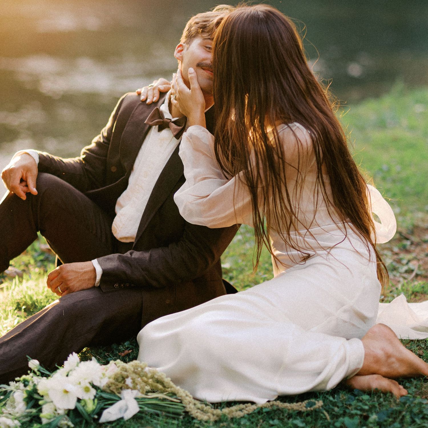 Couple in wedding attire sharing an intimate moment on grass with white flowers.