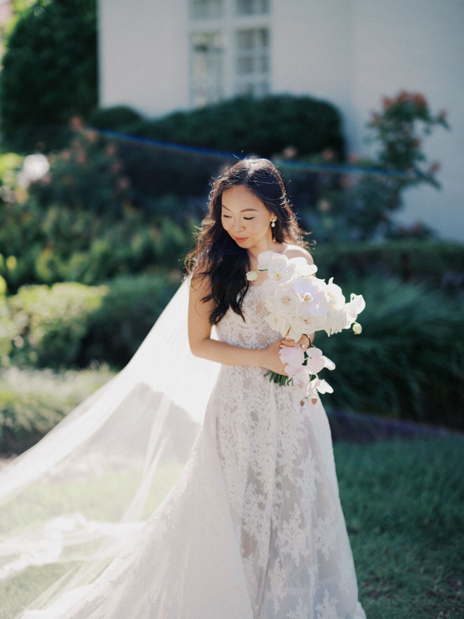 A bride in a delicate lace wedding gown holds a bouquet of white flowers in a sunlit garden setting.