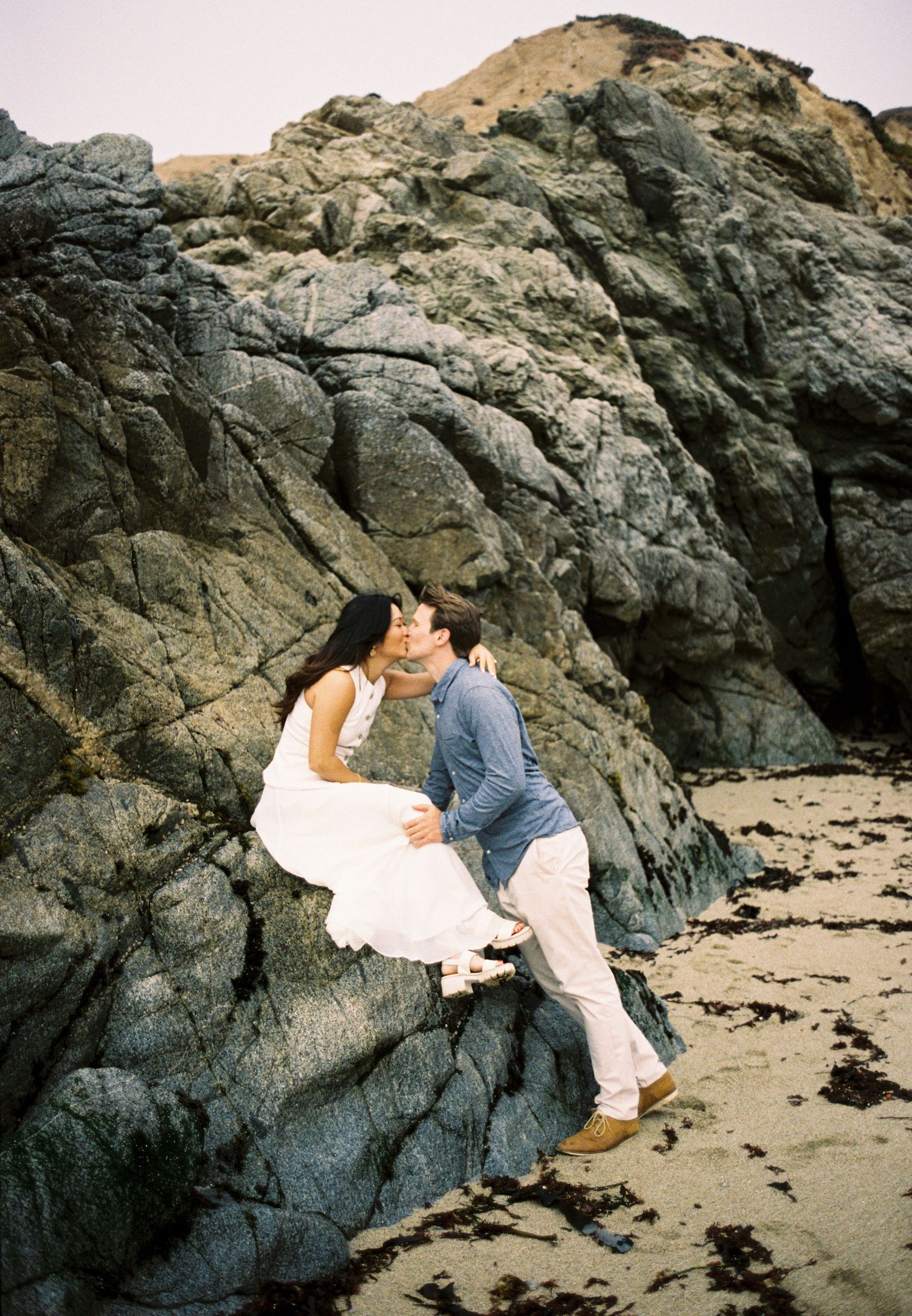 A romantic couple shares tender moments on a rocky beach against dramatic black stone formations at sunset.