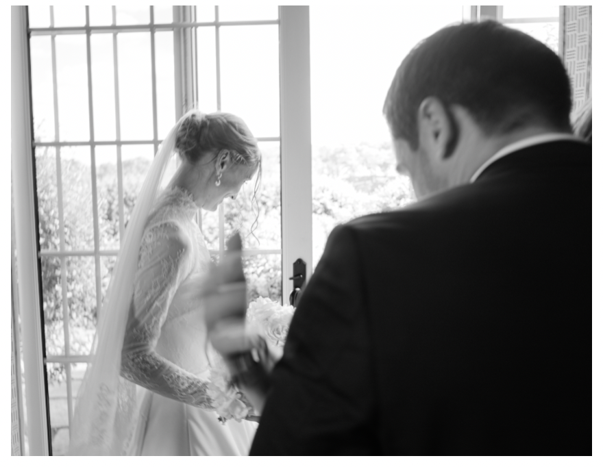 Black and white photo of a bride in a wedding veil holding flowers, facing a groom in a suit near a window.
