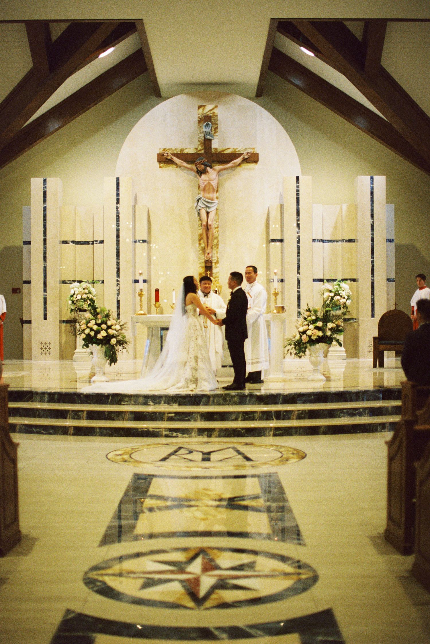 A wedding ceremony takes place at the altar of a Catholic church with ornate floor designs and a large crucifix mounted above.