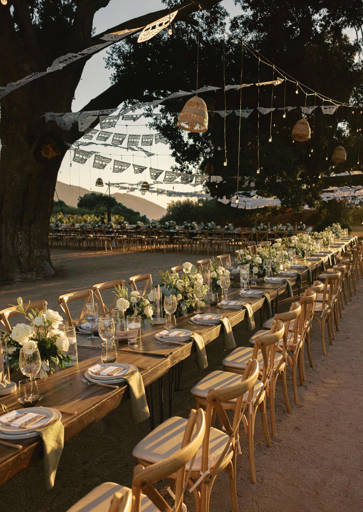 Elegant outdoor wedding reception setup with long wooden tables, string lights, and white floral arrangements under a tent.